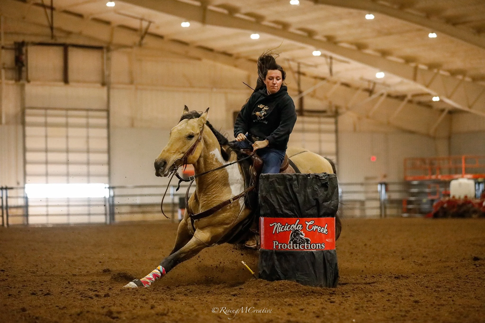 A woman riding a light-colored horse during an indoor equestrian event. The horse is jumping over an obstacle titled 'Nicciola Creek Productions' in a large, well-lit arena with a high ceiling.