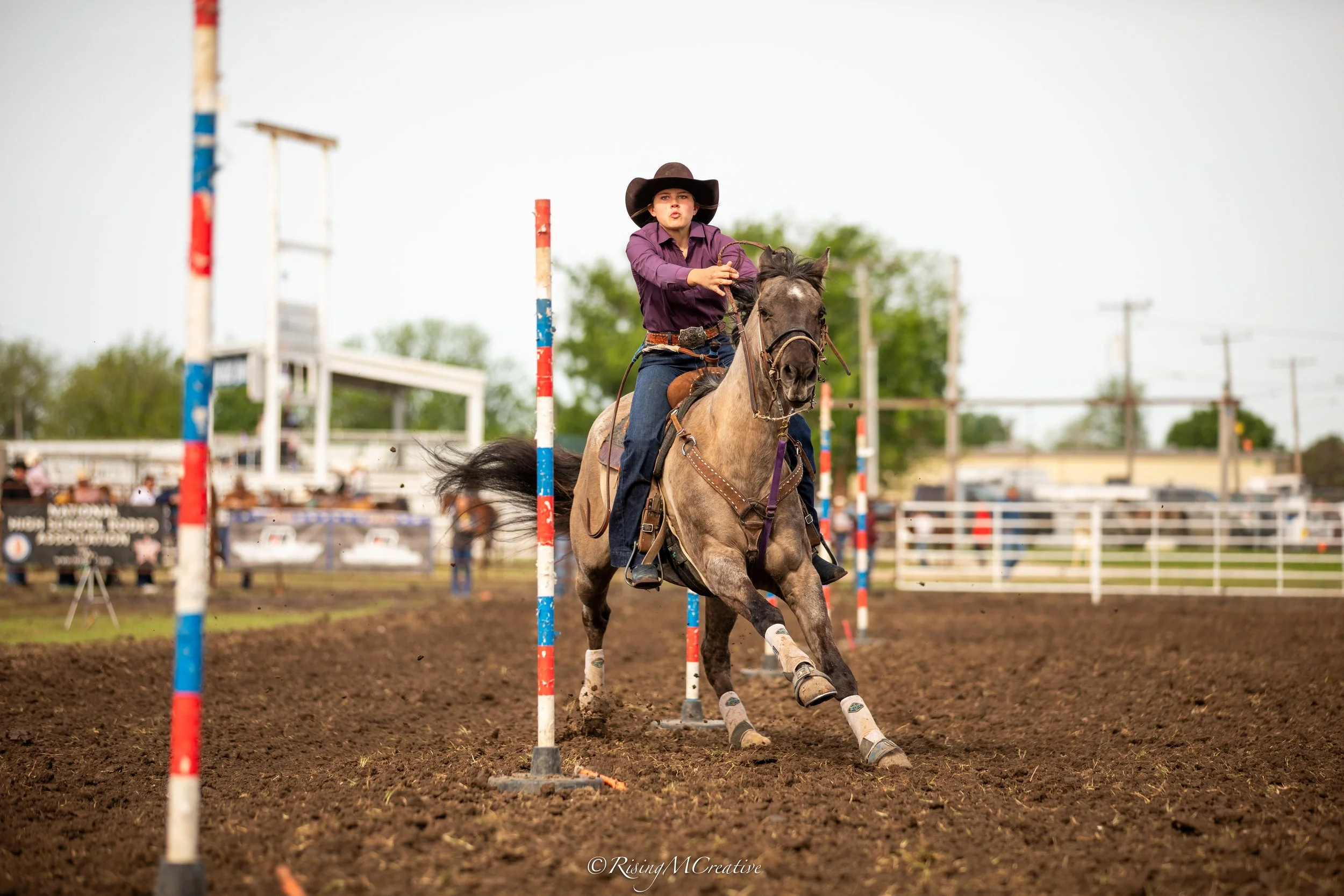 A young woman in a purple shirt and wide-brimmed hat riding a horse and navigating through a slalom course at a rodeo or horse riding event.
