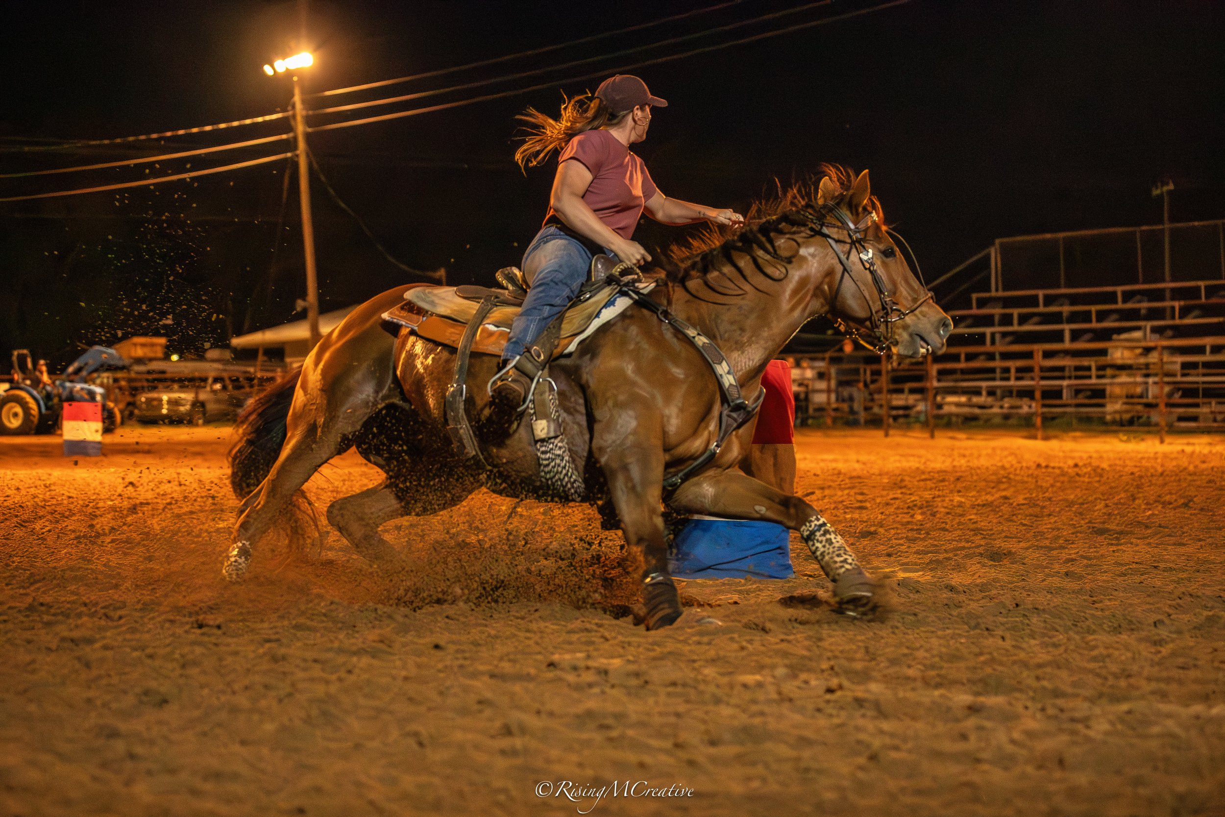 A woman riding a galloping horse at night on a dirt track, with stadium lights and empty bleachers in the background.