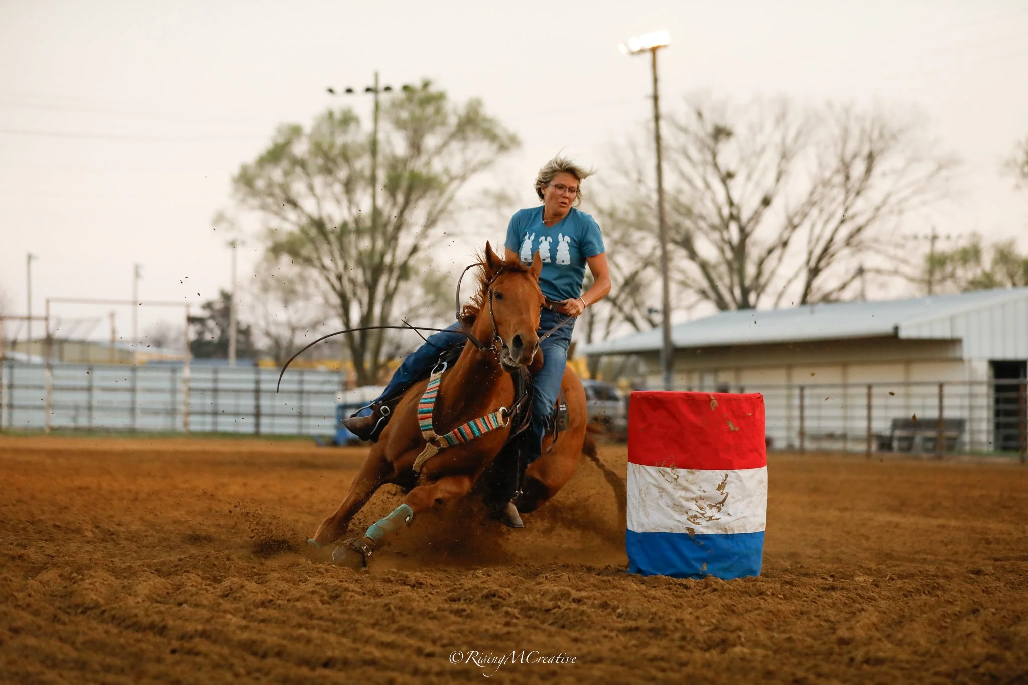 A woman wearing glasses and a blue t-shirt with white rabbits is riding a chestnut horse around a barrel during daytime at a rodeo or equestrian event. The background shows trees, utility poles, and a cloudy sky, with the horse kicking up dirt on an 