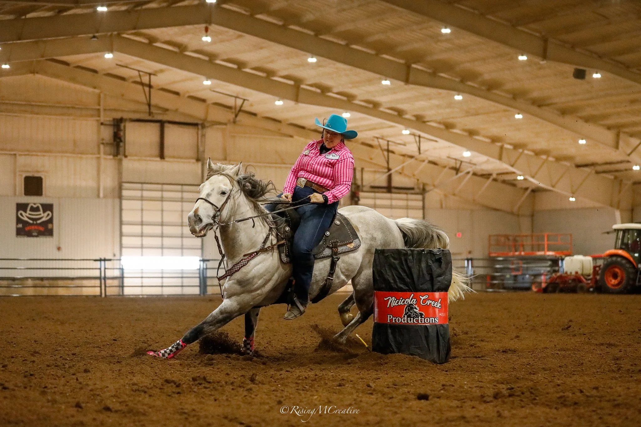 A woman wearing a turquoise cowboy hat and pink checkered shirt riding a gray horse inside an indoor arena, with a barrel labeled Nicciella Creek Productions in front of her, participating in a barrel racing event.