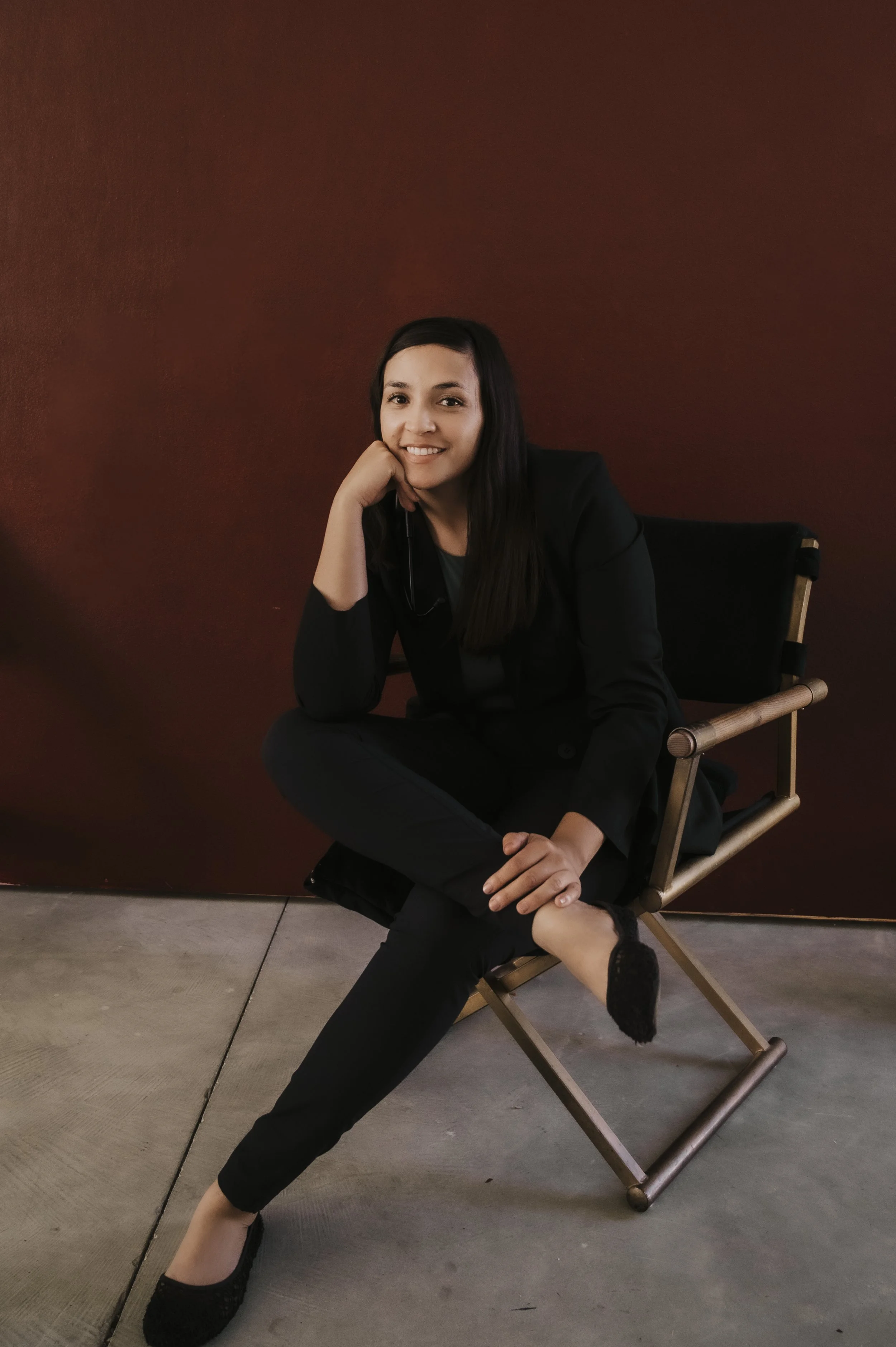 A woman dressed in black sitting cross-legged on a director's chair against a dark red background, smiling with her head resting on her hand.