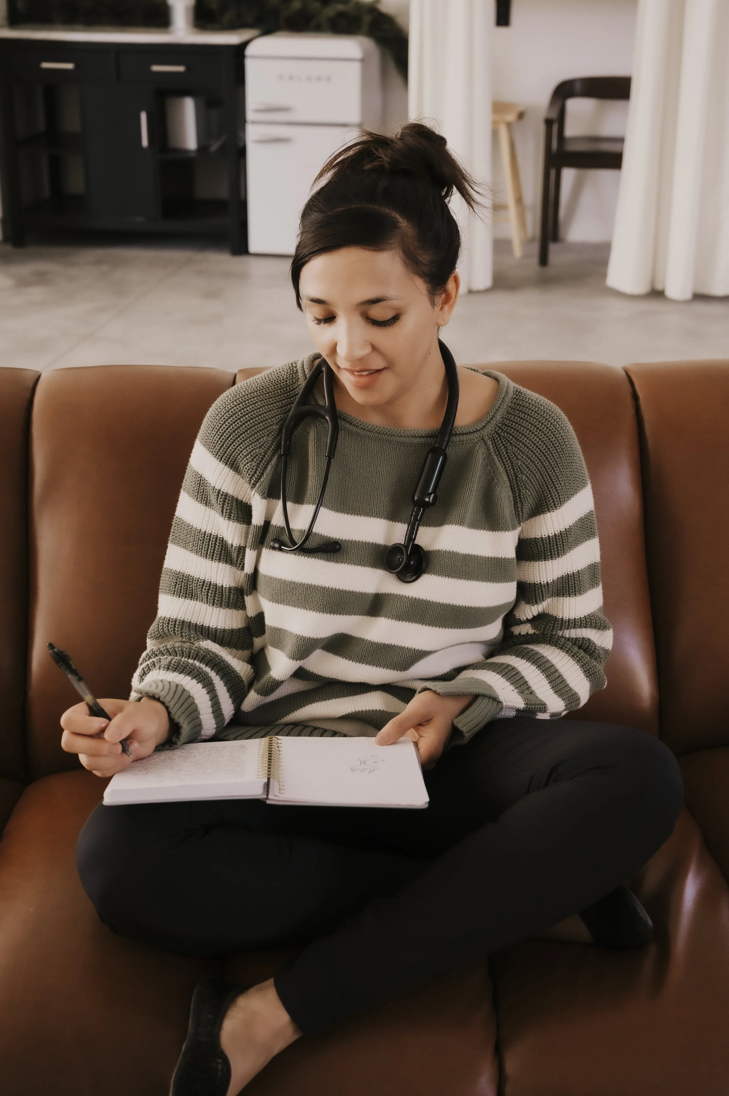 A woman sitting cross-legged on a brown leather couch in a cozy room, with a stethoscope around her neck, writing in a notebook.