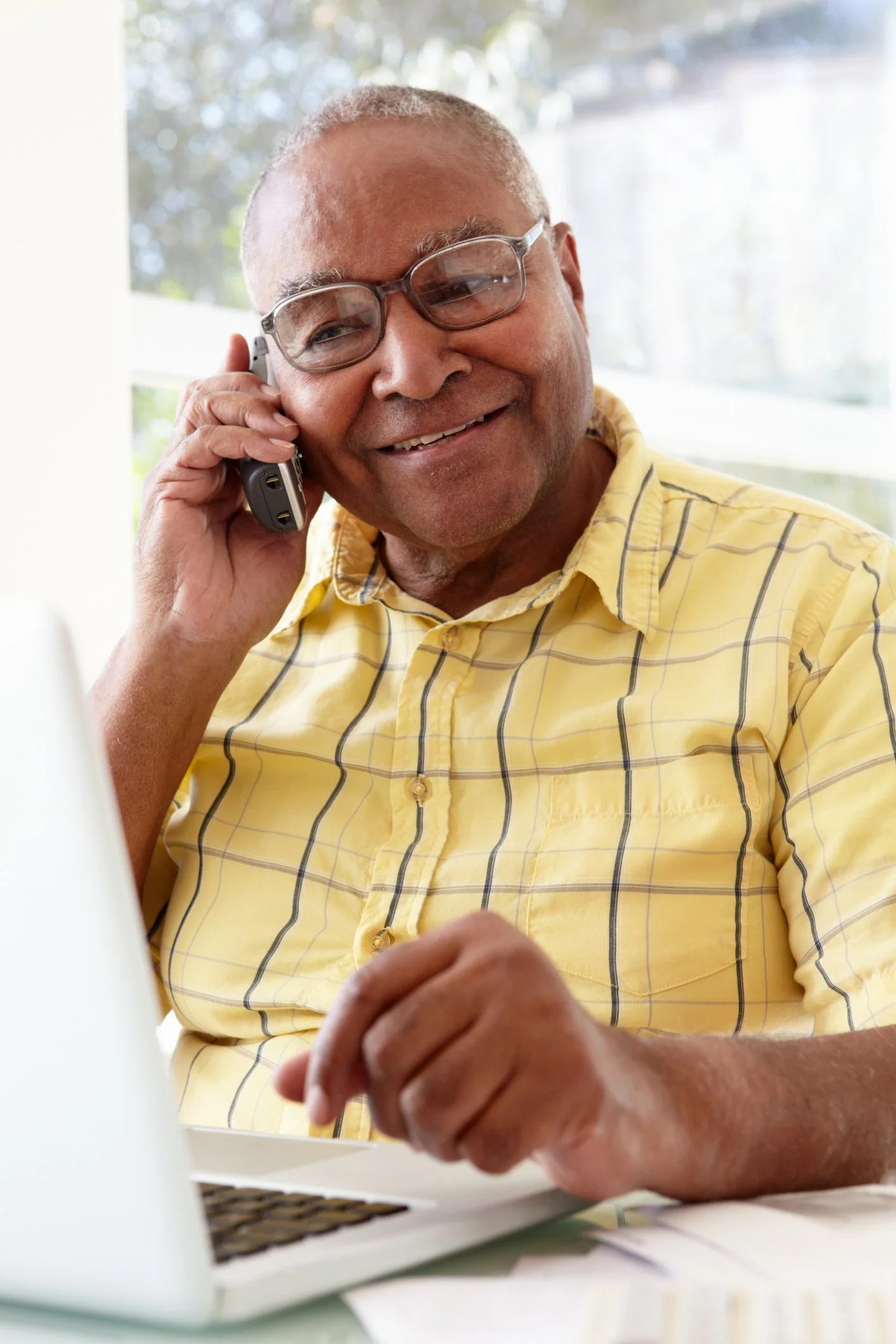 An elderly man with glasses, wearing a yellow checkered shirt, is smiling while talking on a mobile phone and looking at a laptop on a desk.