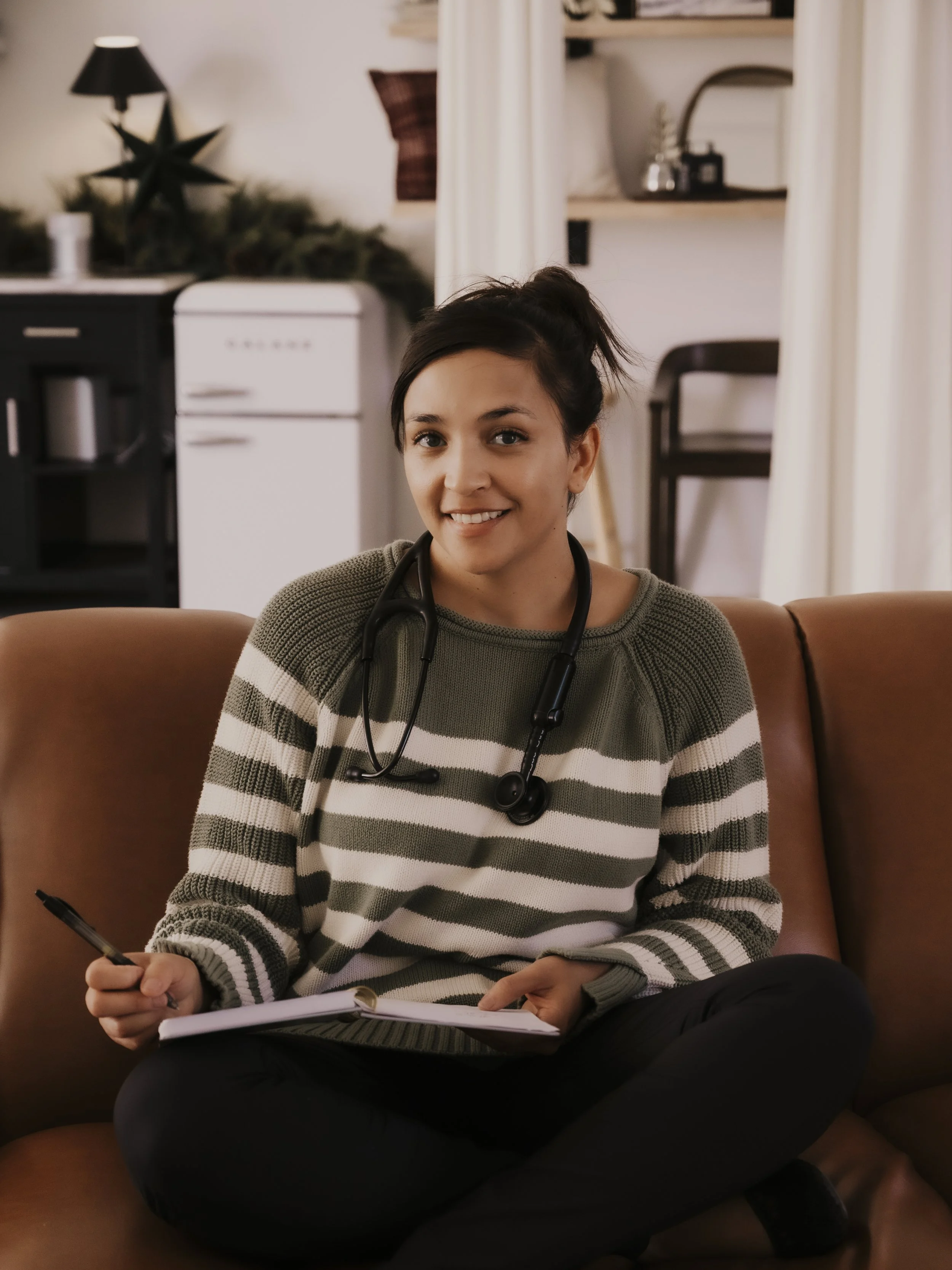 A young woman sitting cross-legged on a couch wearing a striped sweater with a stethoscope around her neck, holding a pen and a notebook, smiling at the camera.