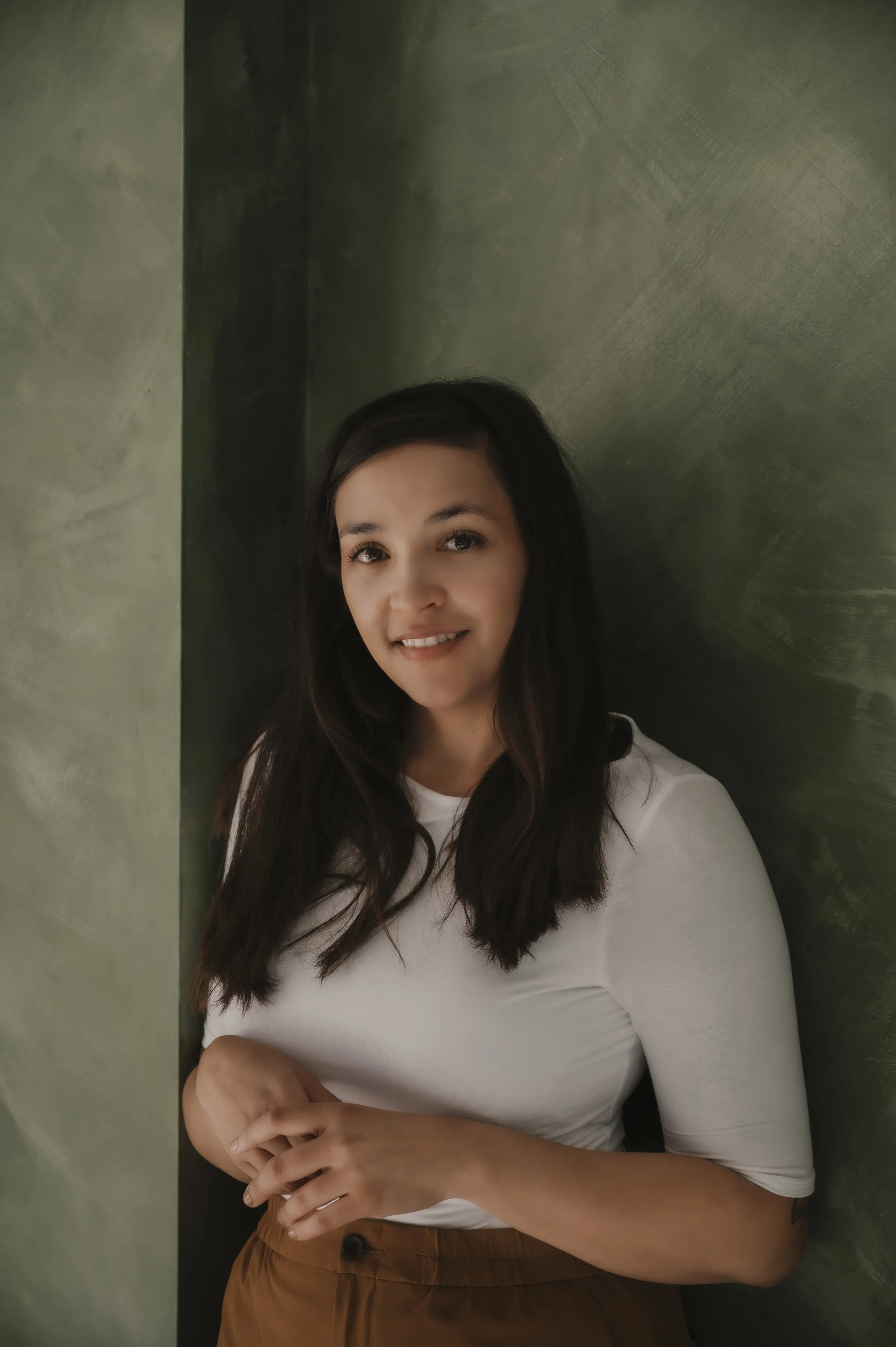 A woman with wavy dark hair smiling, wearing a white top and brown pants, standing against a green textured wall.