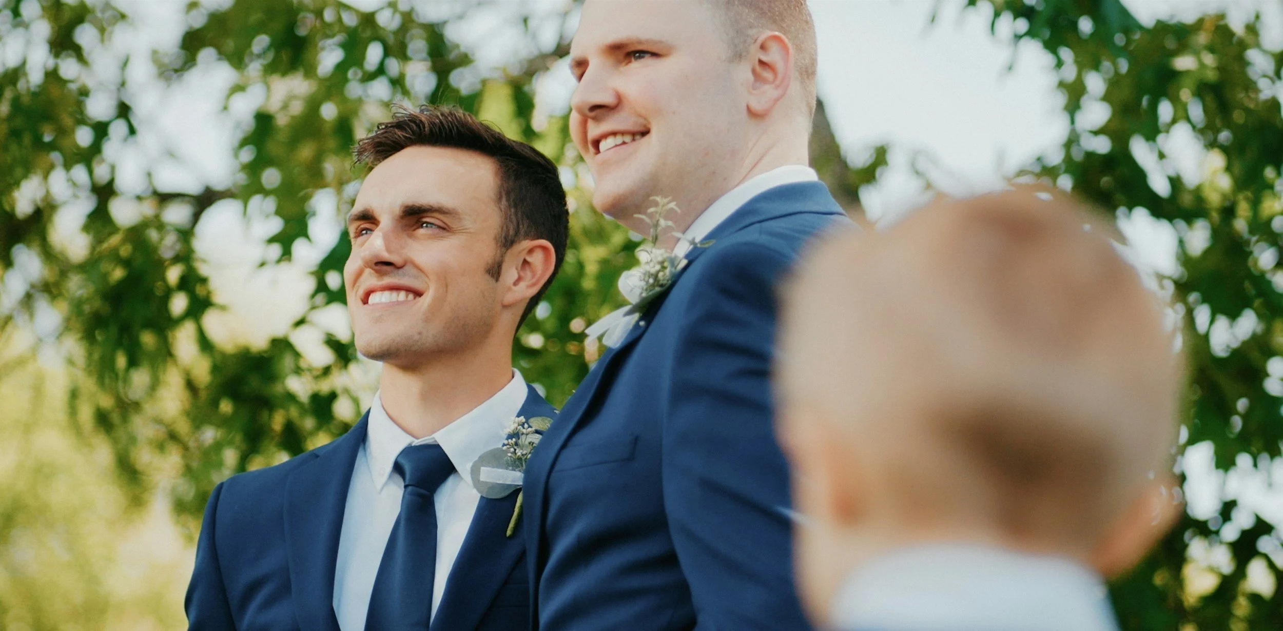Two men in blue suits and ties standing outdoors with green trees in the background, smiling during a wedding ceremony.
