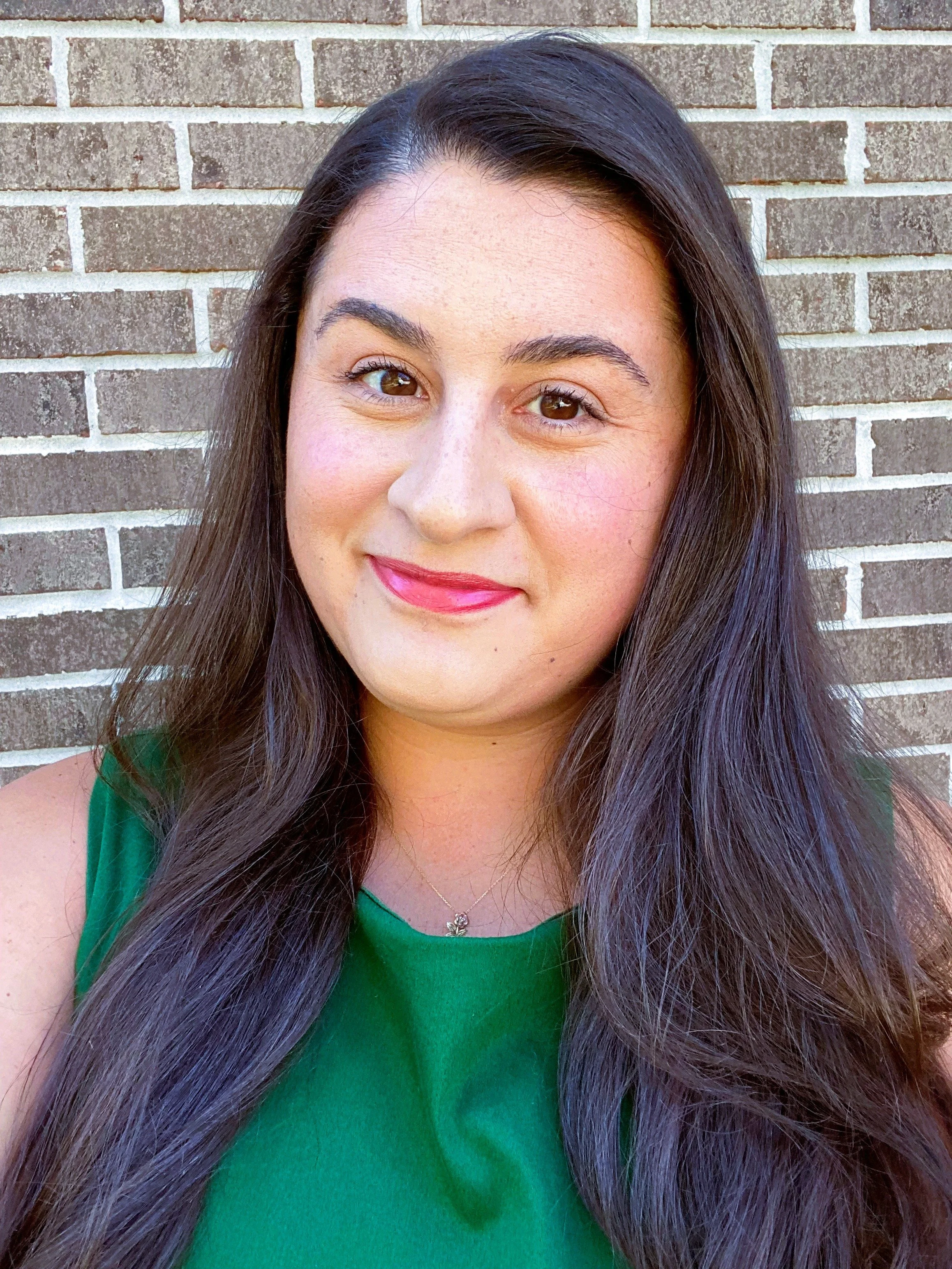 A woman with long dark hair and light skin, smiling, standing in front of a brick wall, wearing a green top and a necklace.