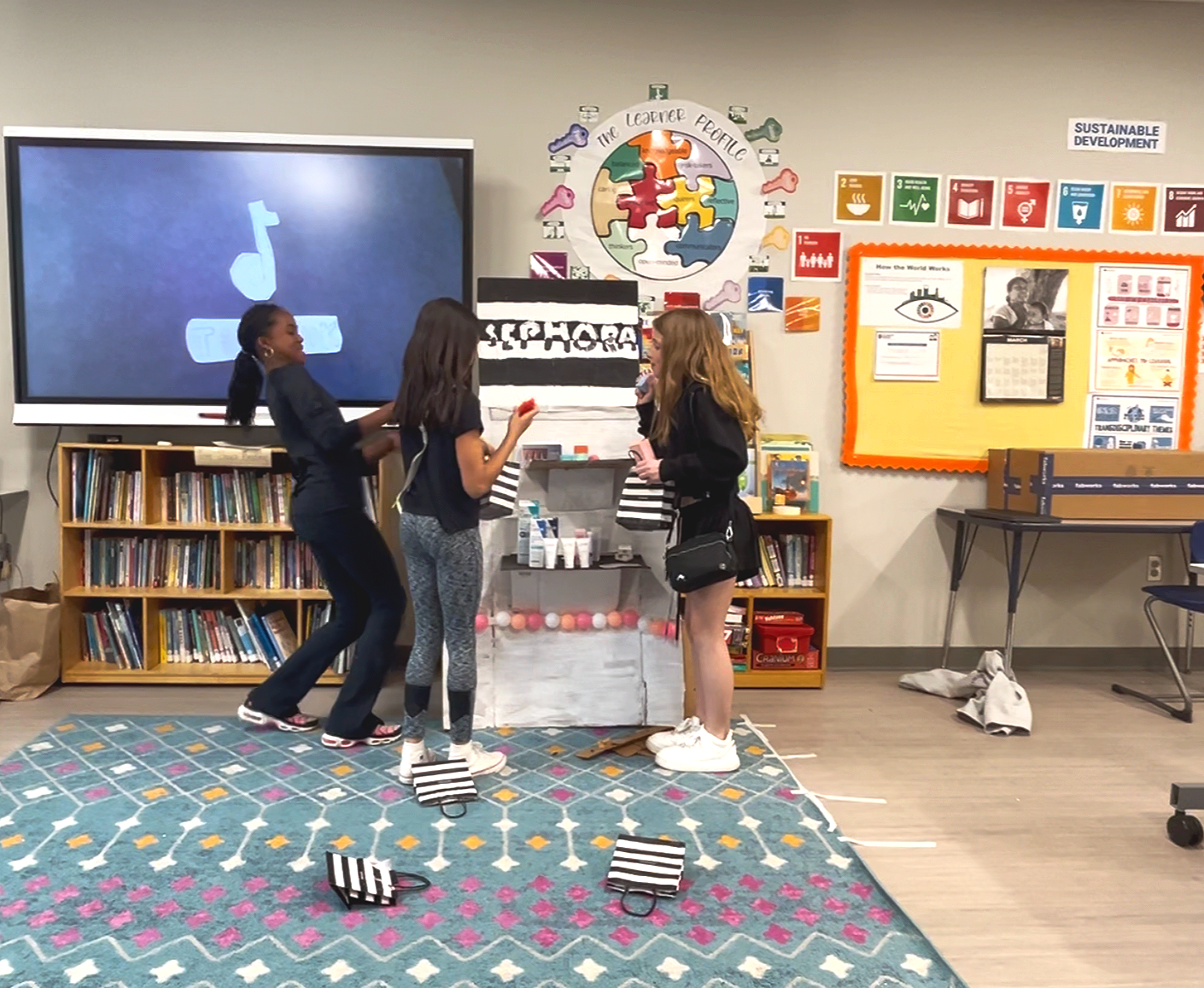 Three girls are in a classroom around a decorated box, appearing to perform a skit or activity. One girl on the left is smiling and the girl on the right is speaking into a microphone. Behind them, a large screen displays a music note icon. The classroom walls have posters about sustainable development and world history, with shelves of books and classroom supplies visible.
