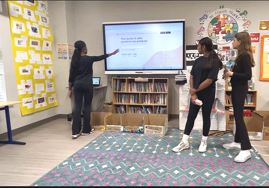 Three young women stand in a classroom, listening to a woman giving a presentation at a digital screen. The room has colorful decorations, bookshelves, and boxes on the floor.