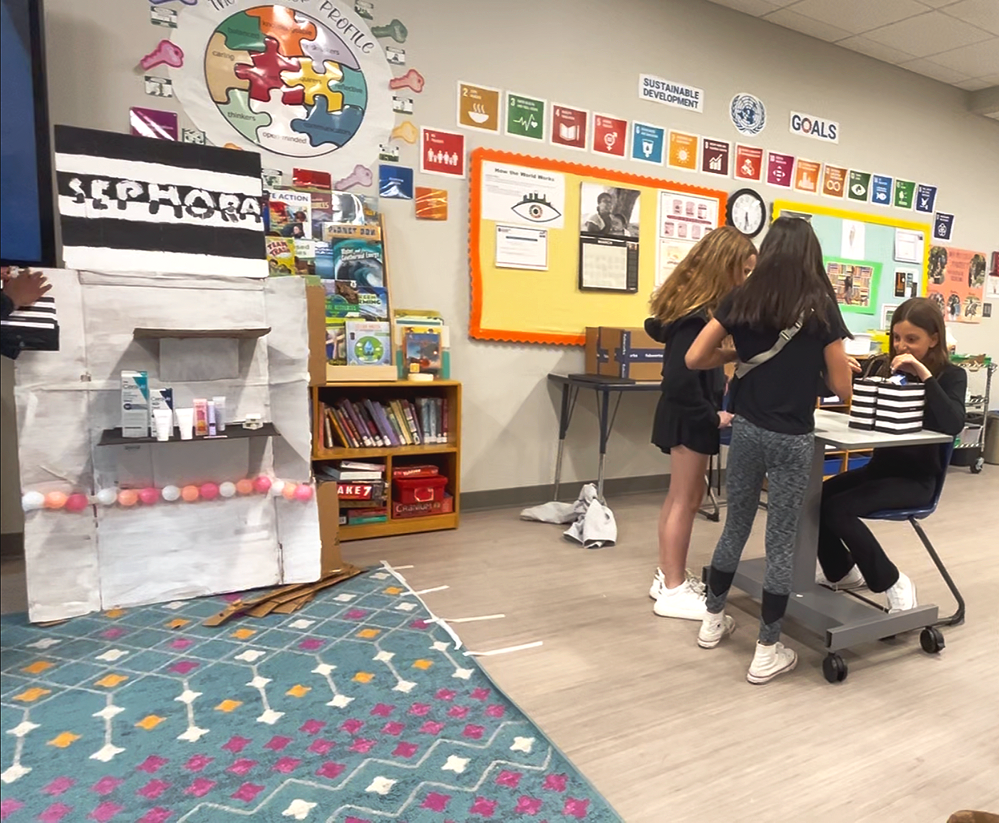 Three girls in black shirts and leggings in a classroom, around a small table with books and supplies, engaging in an activity. A decorated cardboard structure labeled 'SOPHORA' is next to a bookshelf with books and educational materials.