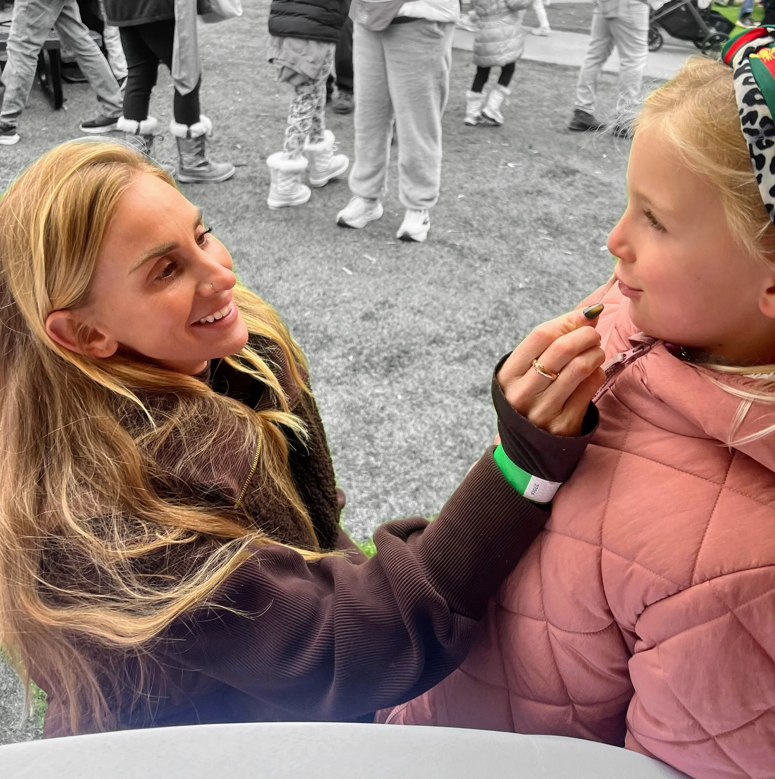 A smiling woman with long blonde hair and a nose piercing is talking into the ear of a young girl with blonde hair, who is wearing a pink quilted jacket and a colorful headband, during an outdoor event. The background shows people walking around on a paved surface, with some in casual winter clothing.