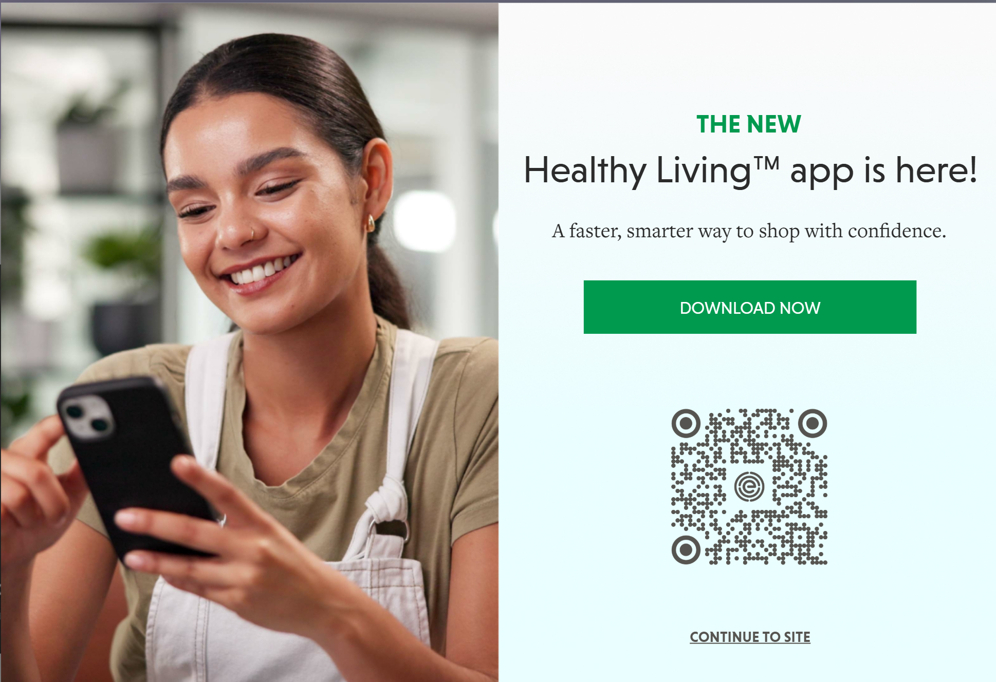 A smiling young woman with dark hair is looking at her phone, wearing a tan shirt and white apron, in a bright indoor space with plants in the background.