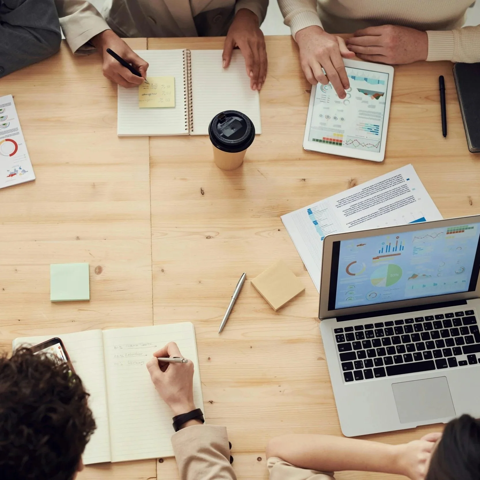 A group of people sitting around a wooden table working on laptops, notebooks, and tablets with charts and data. A takeaway coffee cup is in the center of the table.