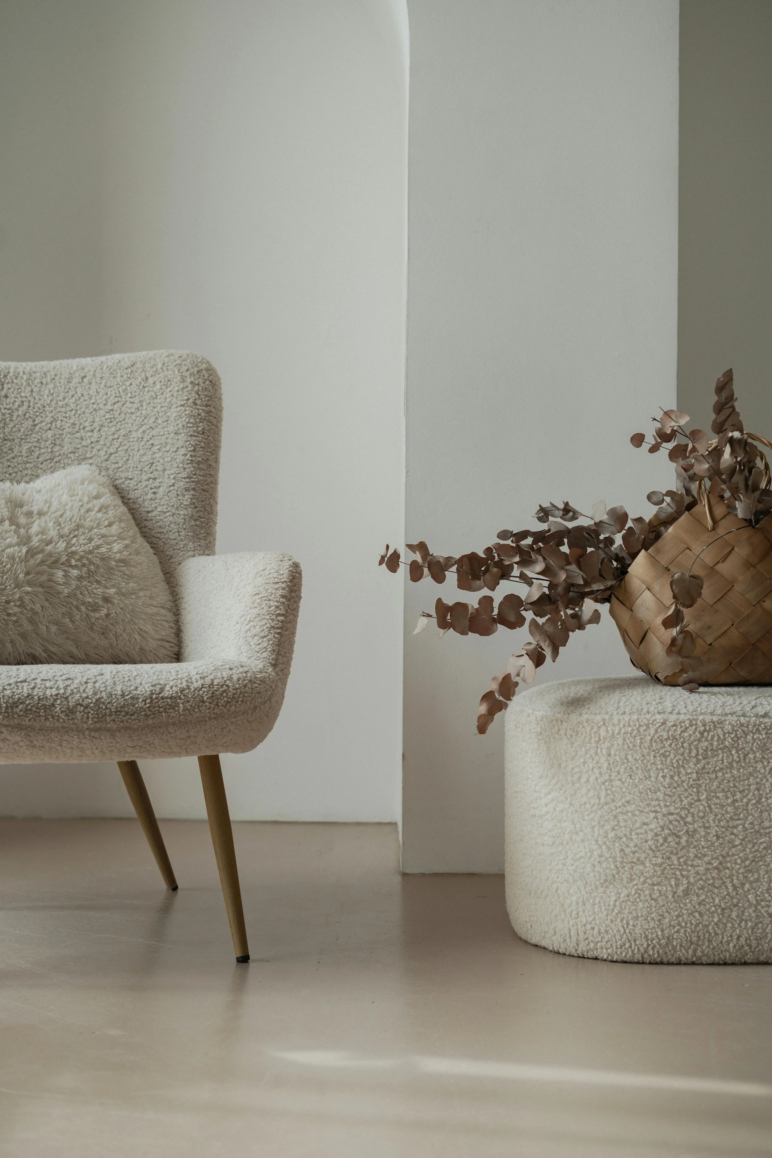 Minimalist interior with a beige armchair, a plush pillow, a woven basket with dried eucalyptus, and simple white walls.