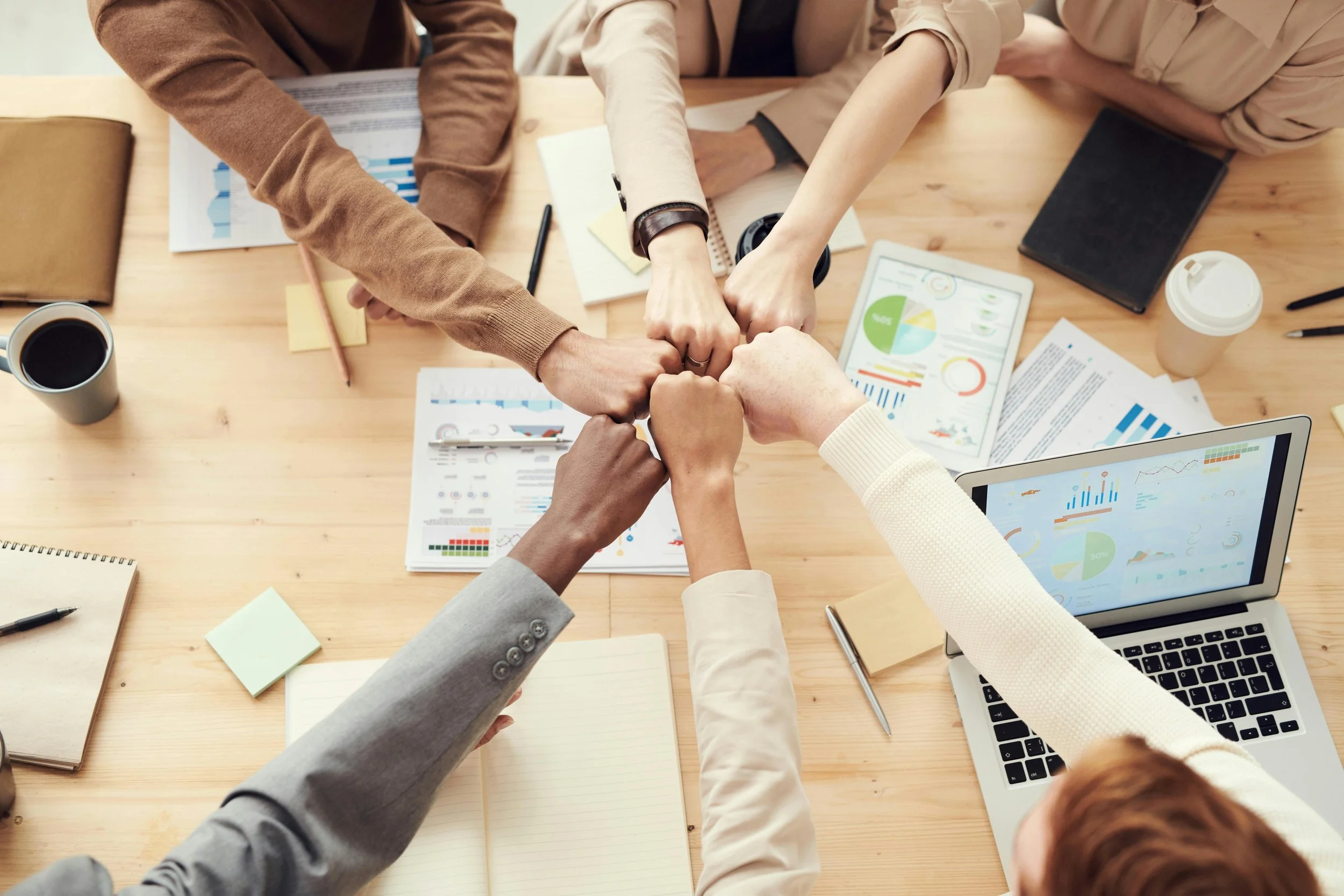 Multiple people putting their fists together in a team huddle over a wooden table with documents, laptops, notebooks, and coffee cups.