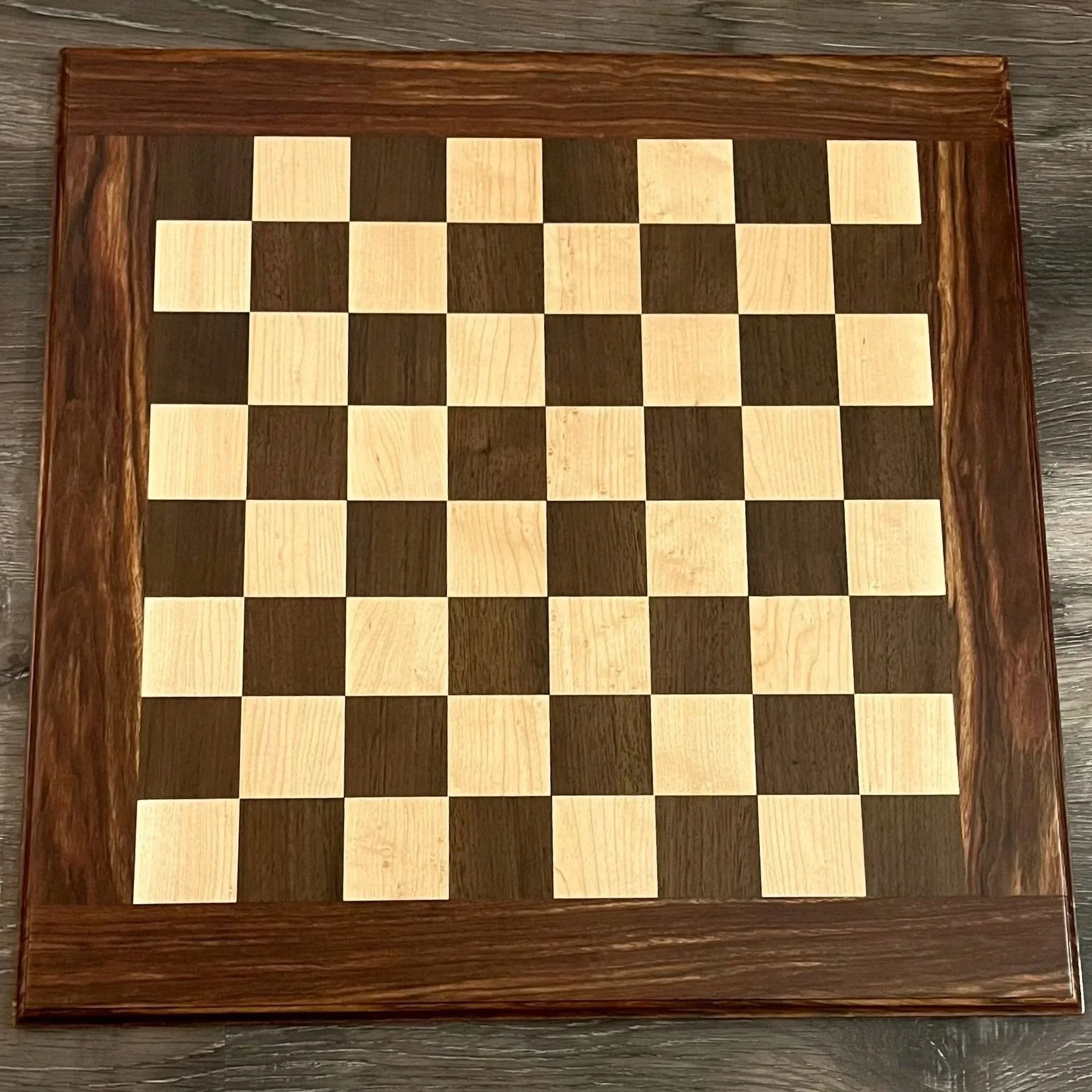 A wooden chessboard with alternating light and dark squares, framed by a dark red rosewood border, placed on a gray wooden surface.