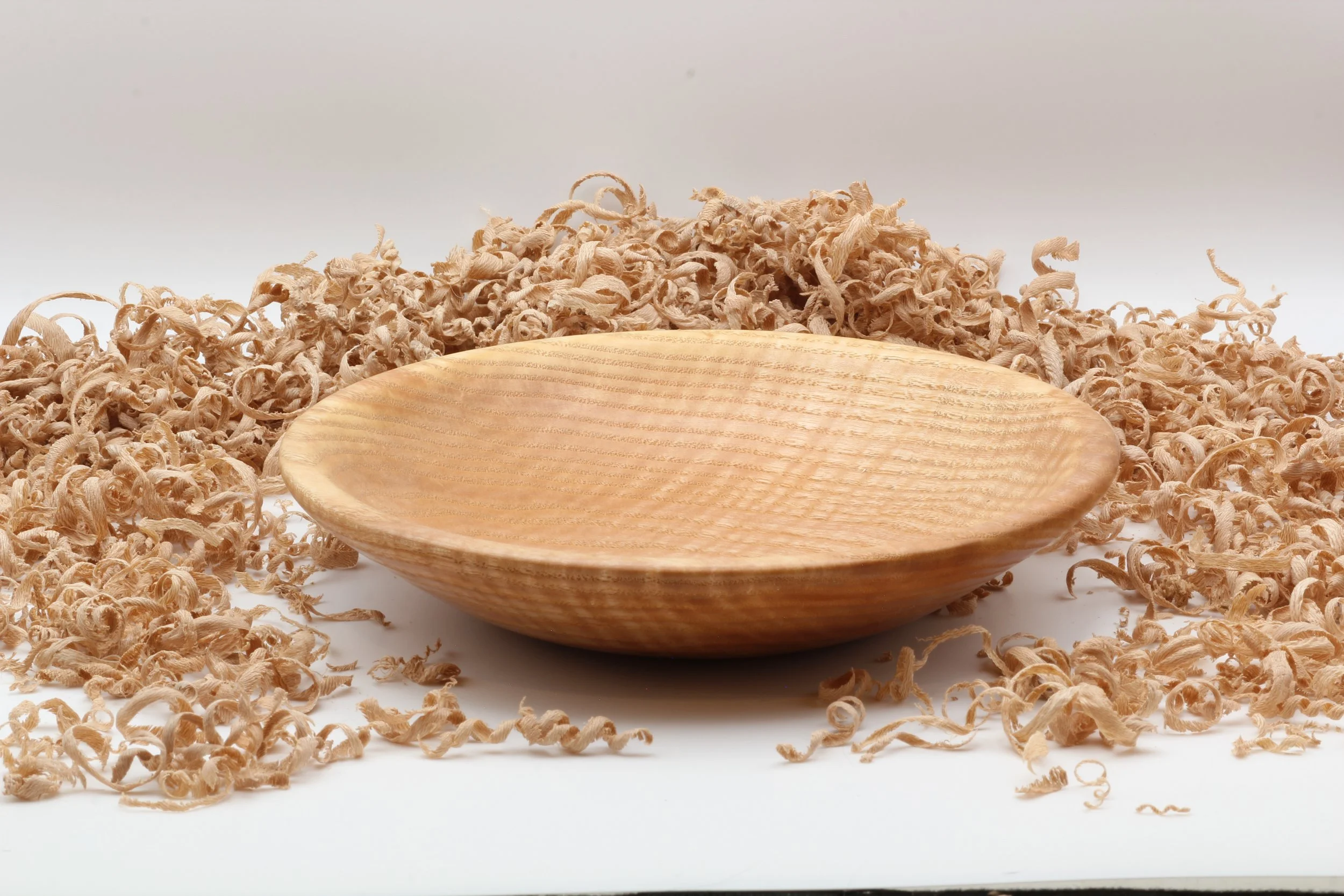 A wooden bowl resting on a surface surrounded by wood shavings, with a plain white background.