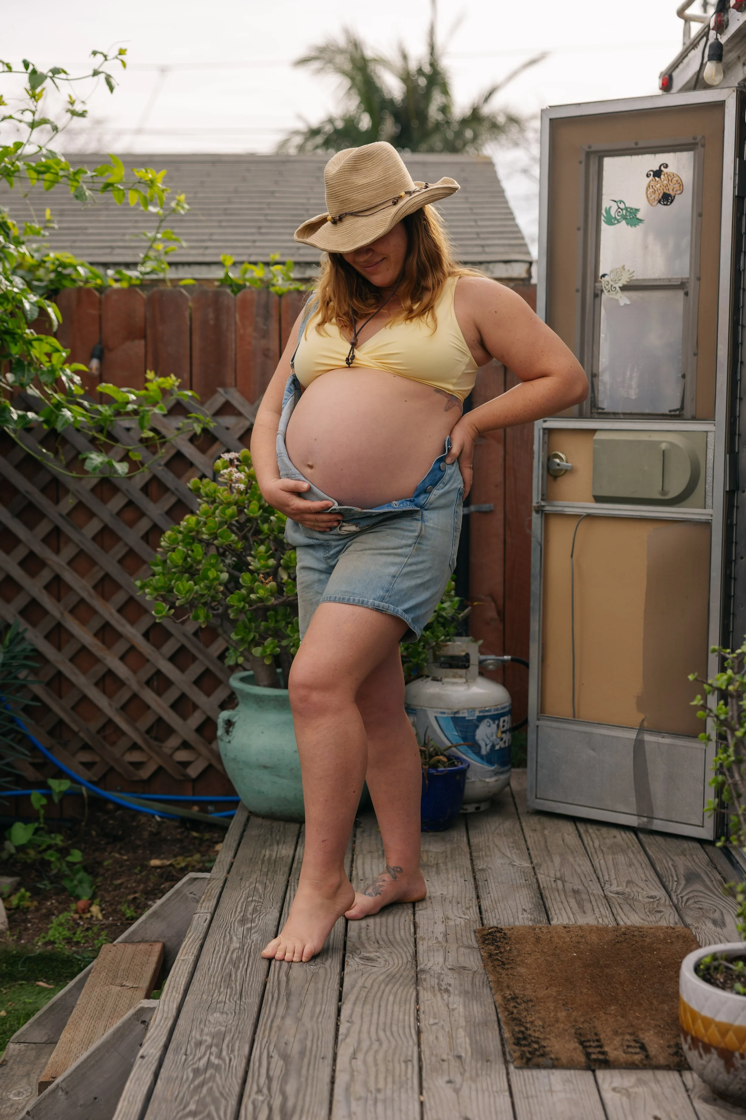 A pregnant woman stands on a wooden deck outdoors, wearing a wide-brimmed hat, yellow top, and denim shorts, smiling and holding her belly.