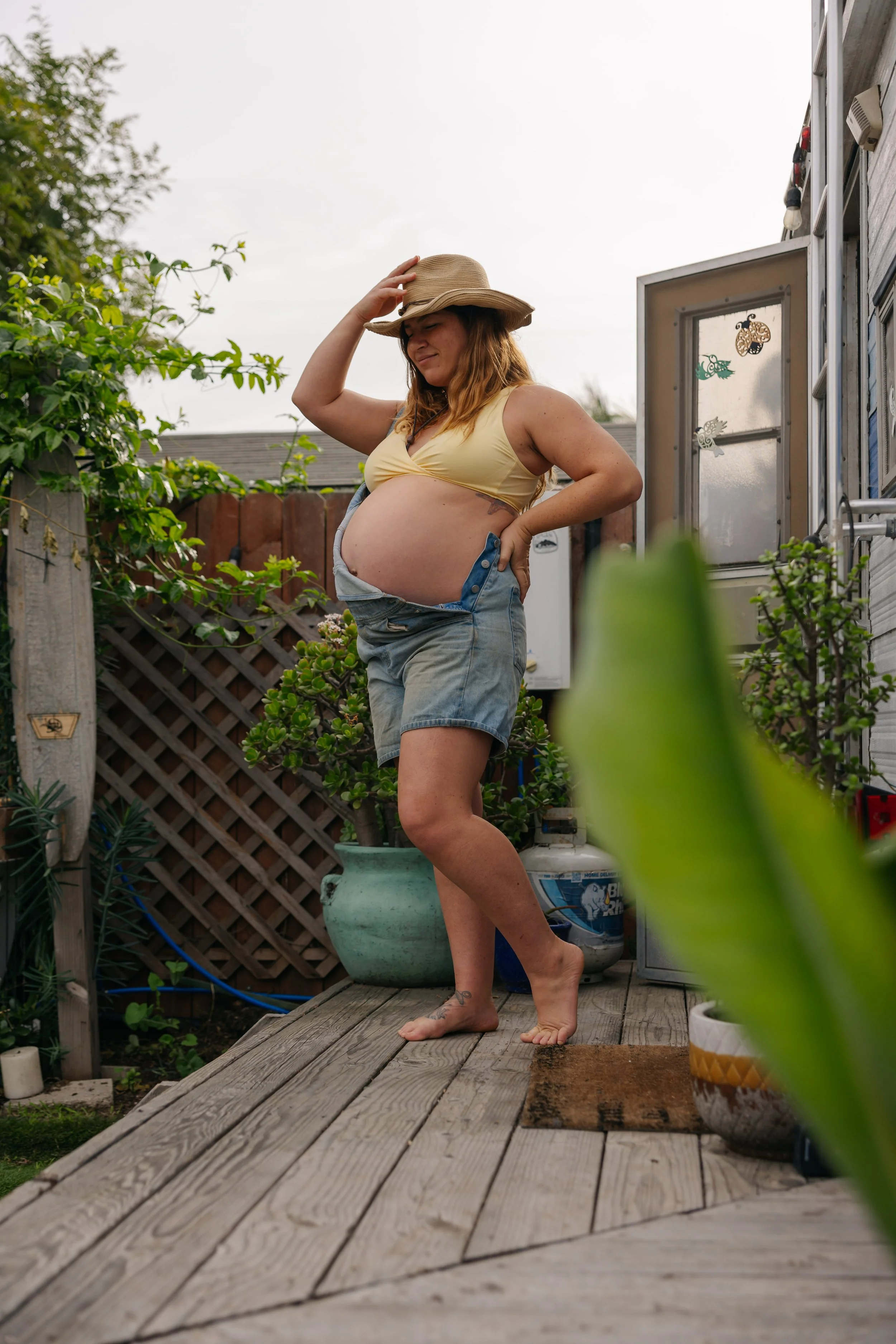 A pregnant woman standing on a wooden deck outdoors, wearing a yellow top, denim shorts, and a straw hat, touching her hat with one hand and resting her other hand on her hip, with greenery and plants around her.