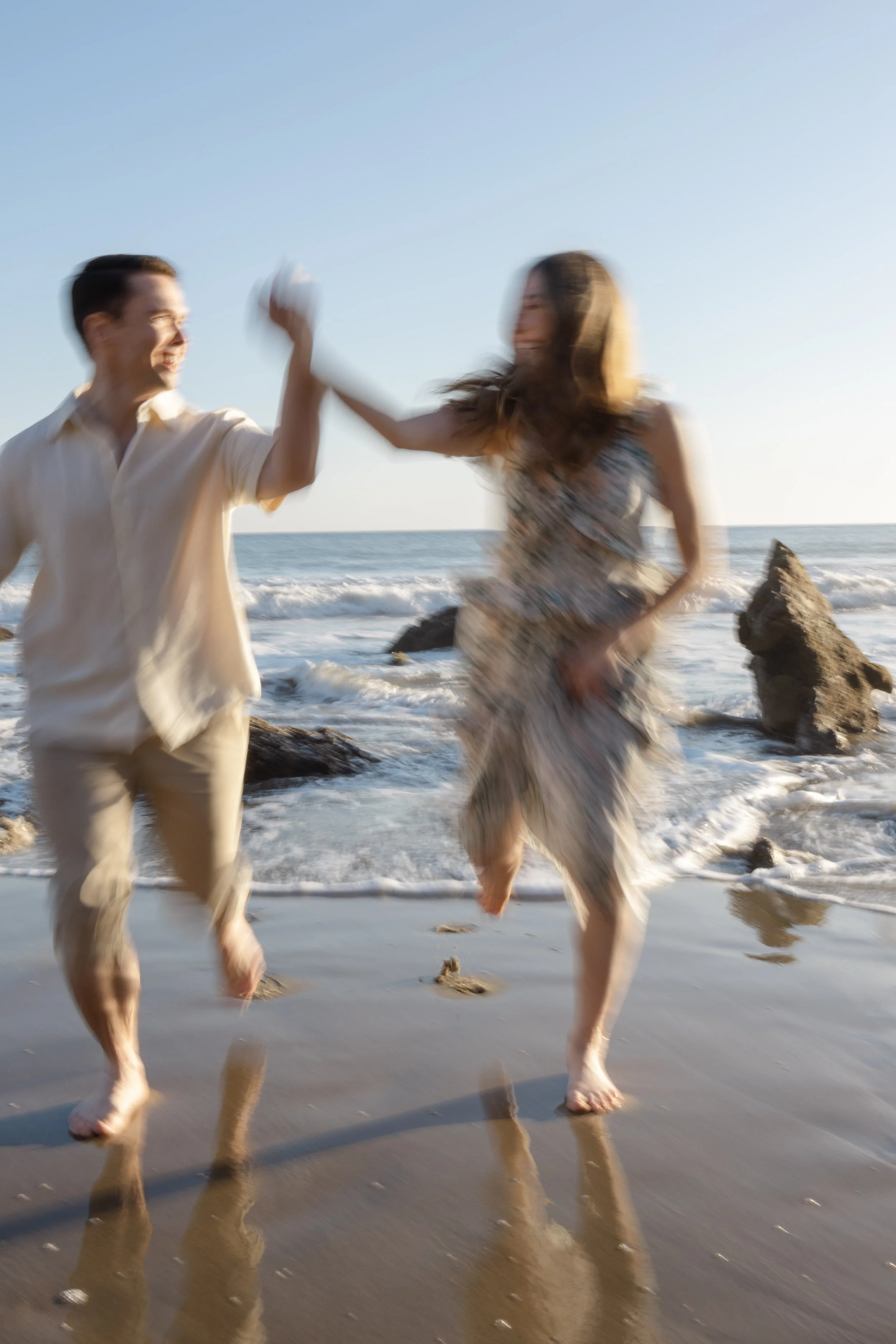 Two people playing on the beach, high-fiving, with ocean waves and rocks in the background.