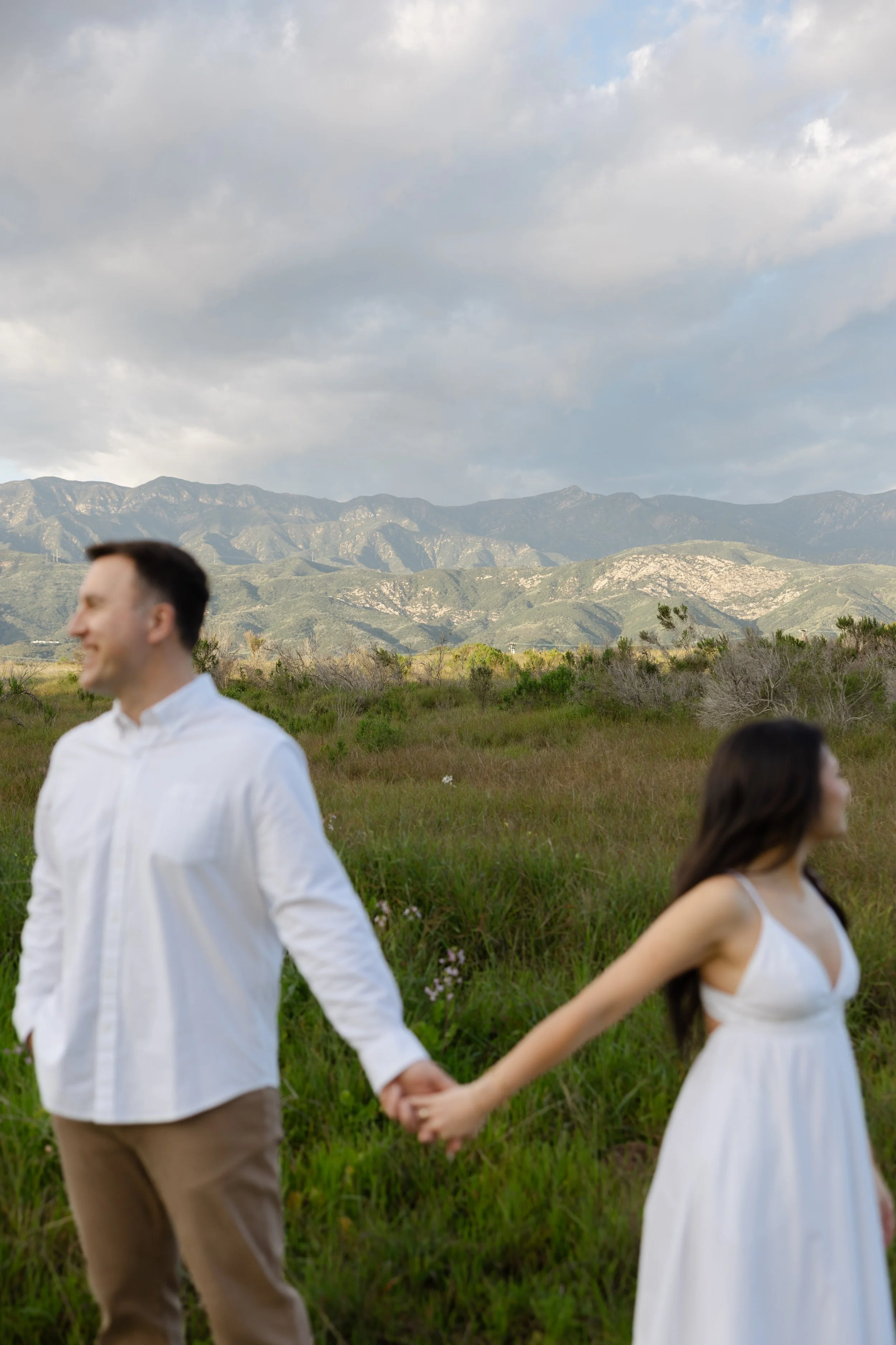 A man and woman holding hands in a grassy field with mountains in the background, under a partly cloudy sky.