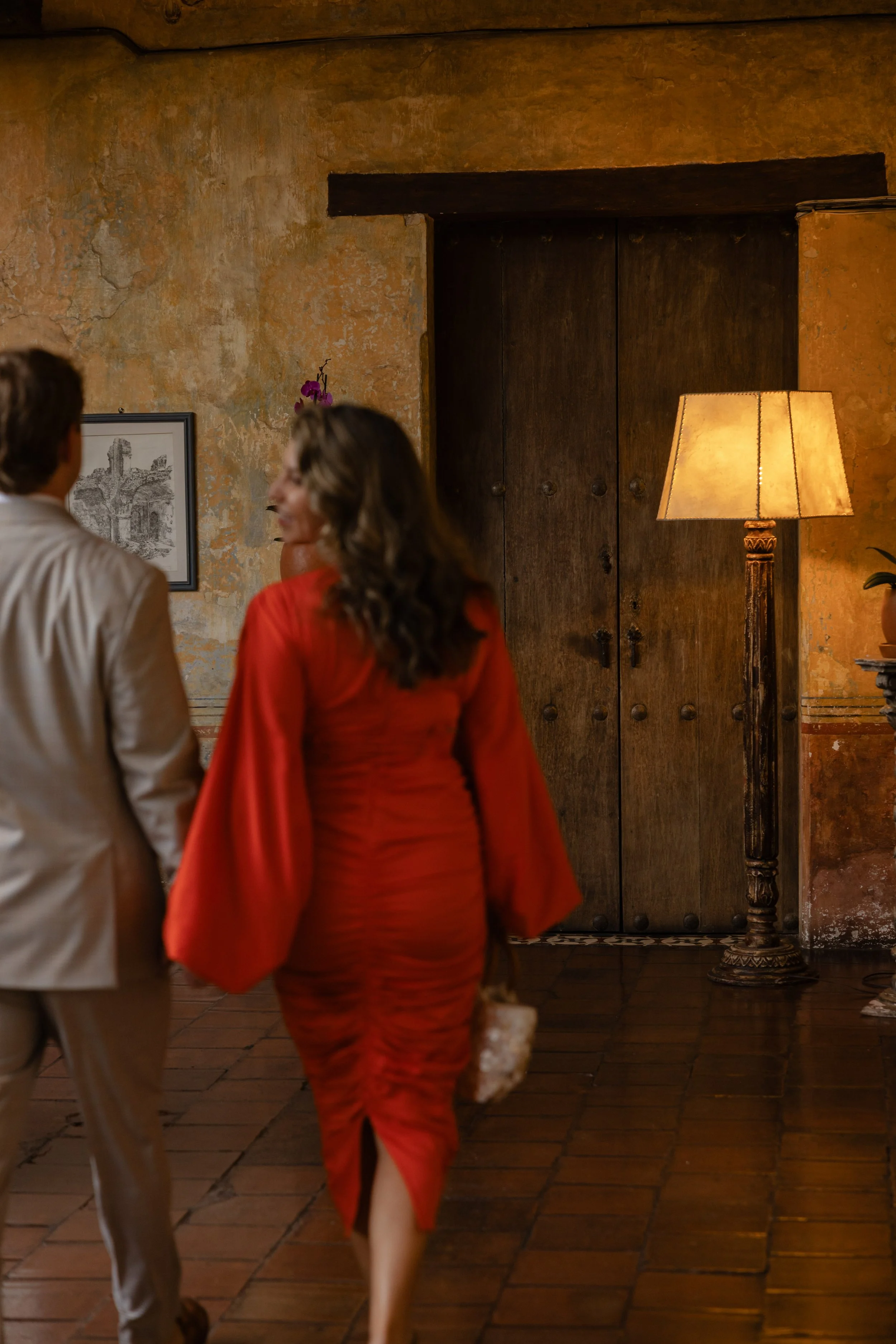 A man and a woman walking and talking inside a warmly lit room with rustic decor, a large wooden door, and a tall standing lamp.
