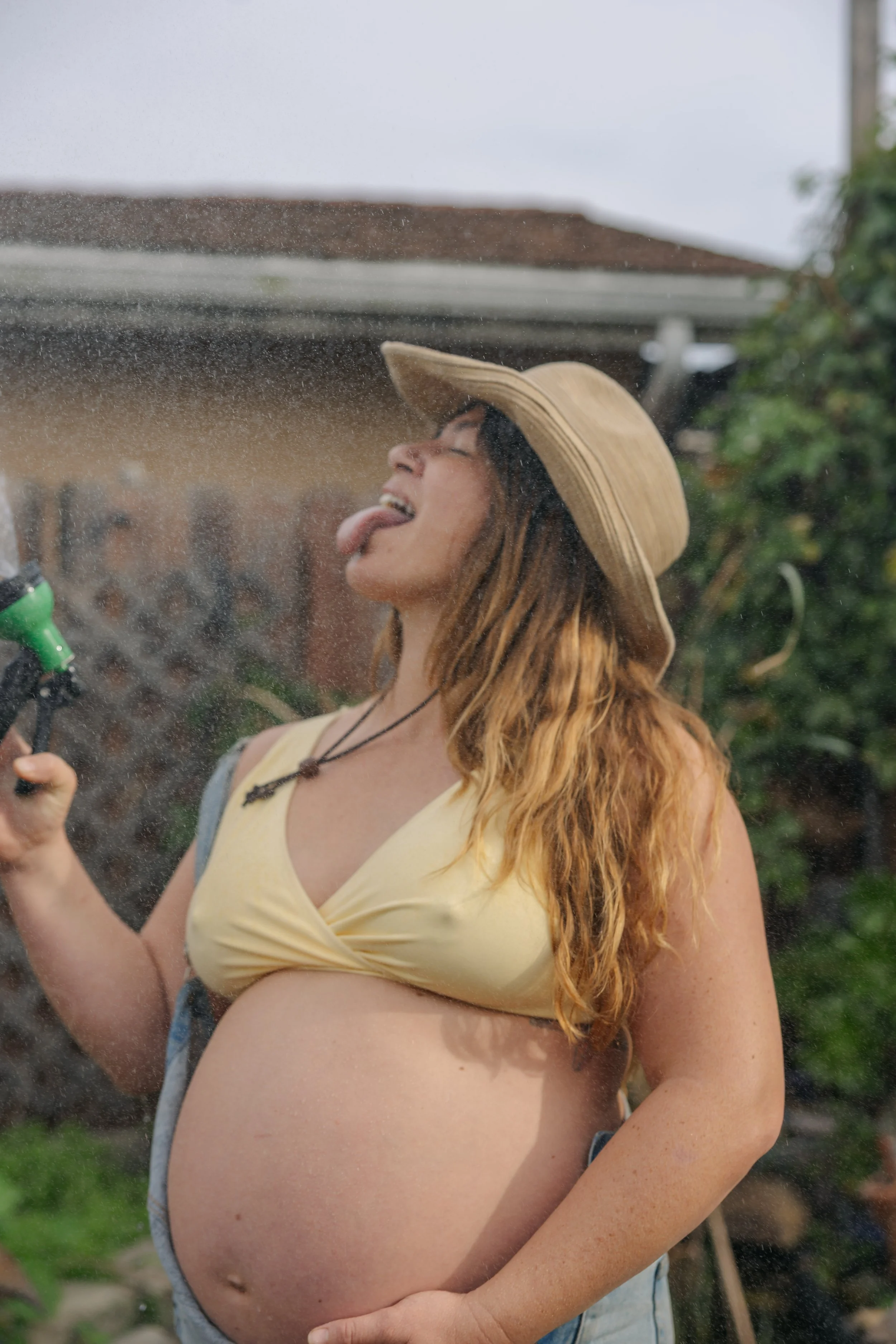 Pregnant woman with long red hair wearing a straw hat and yellow top, sticking out her tongue while watering plants outside on a cloudy day.