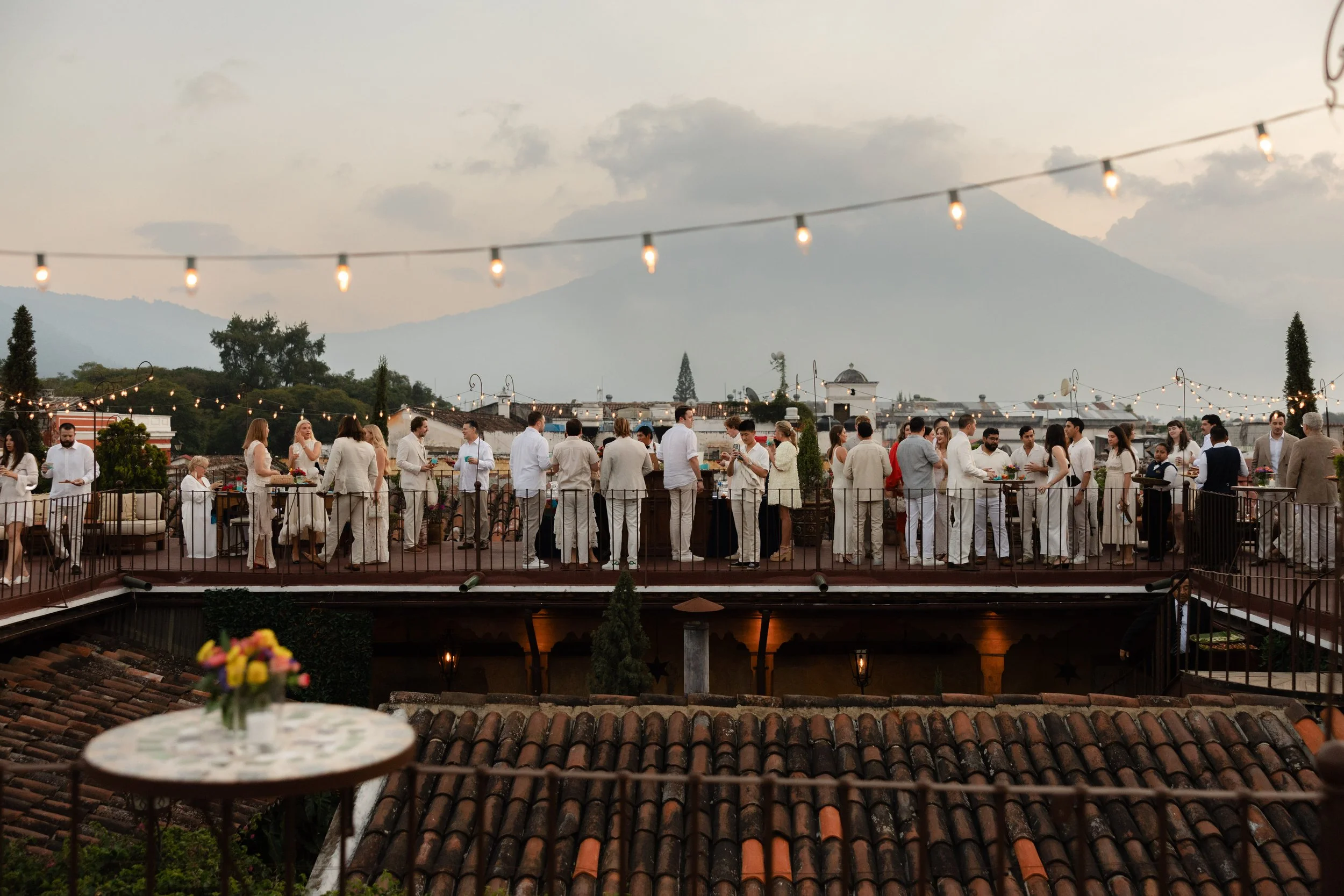 People dressed in white and light-colored clothing celebrating on a rooftop terrace at sunset, with string lights overhead, overlooking a cityscape and mountains in the background.