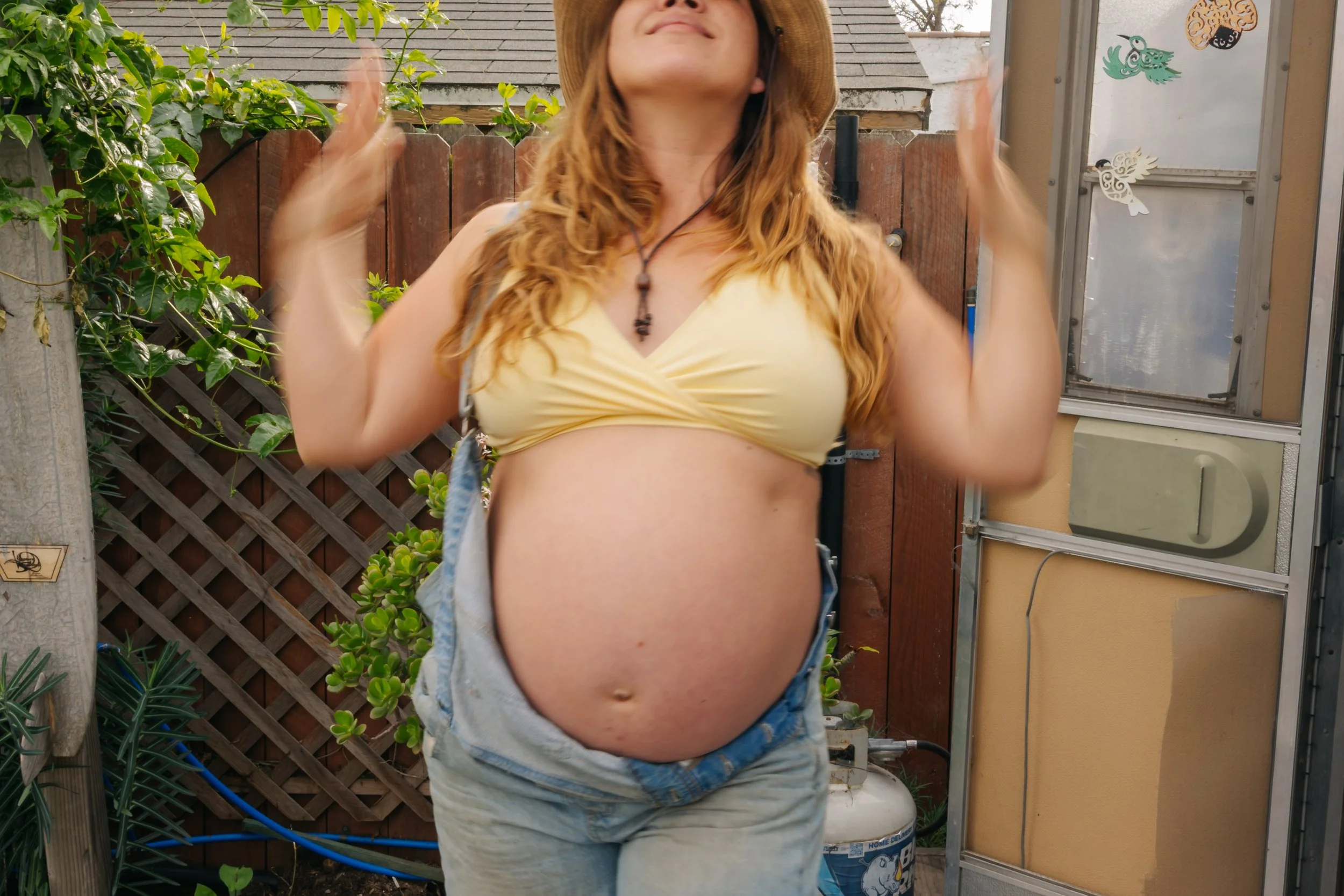 A pregnant woman with red hair wearing a yellow top, blue jeans, and a large straw hat standing outdoors in a garden.