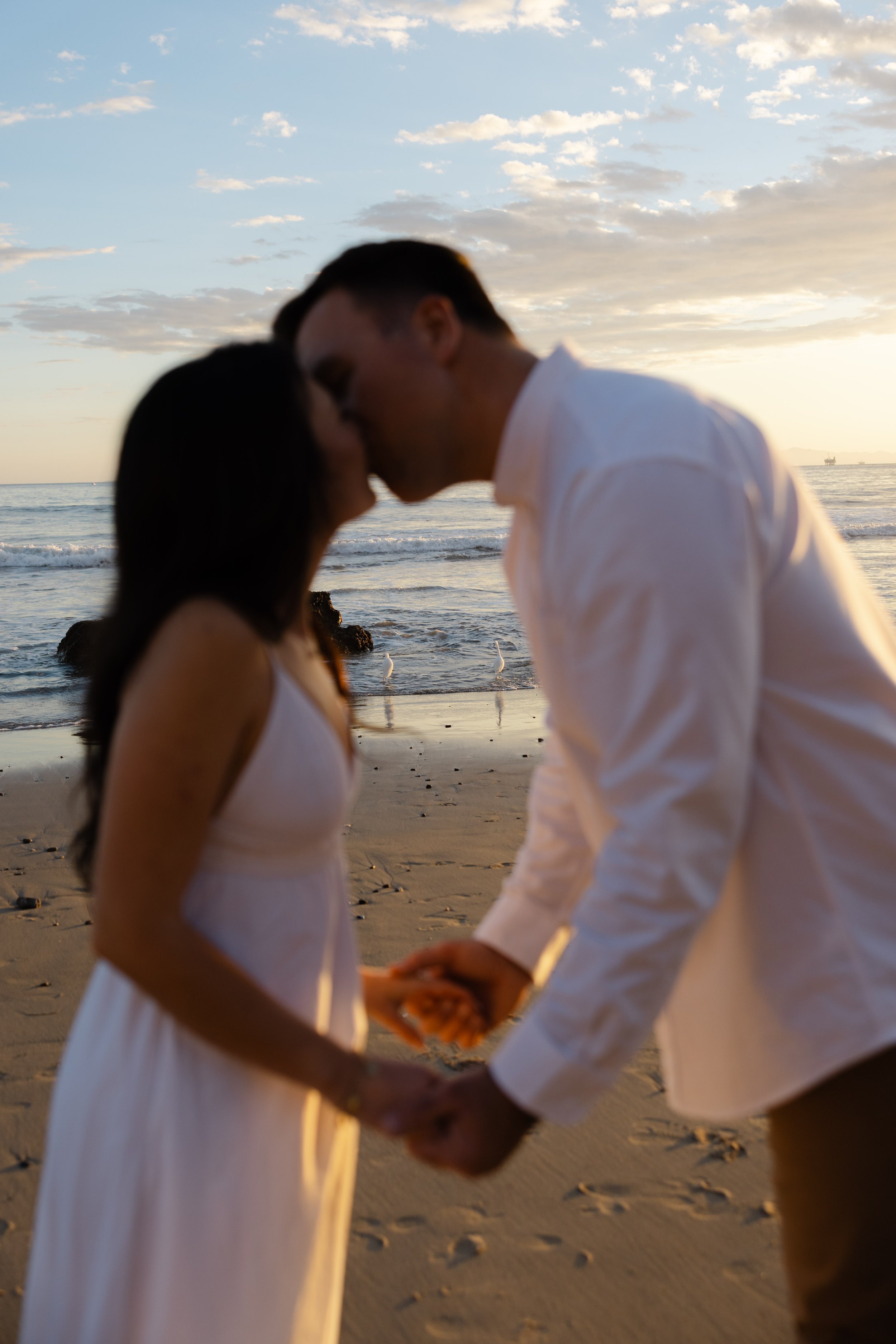 A couple in white clothing holding hands and kissing on the beach during sunset.