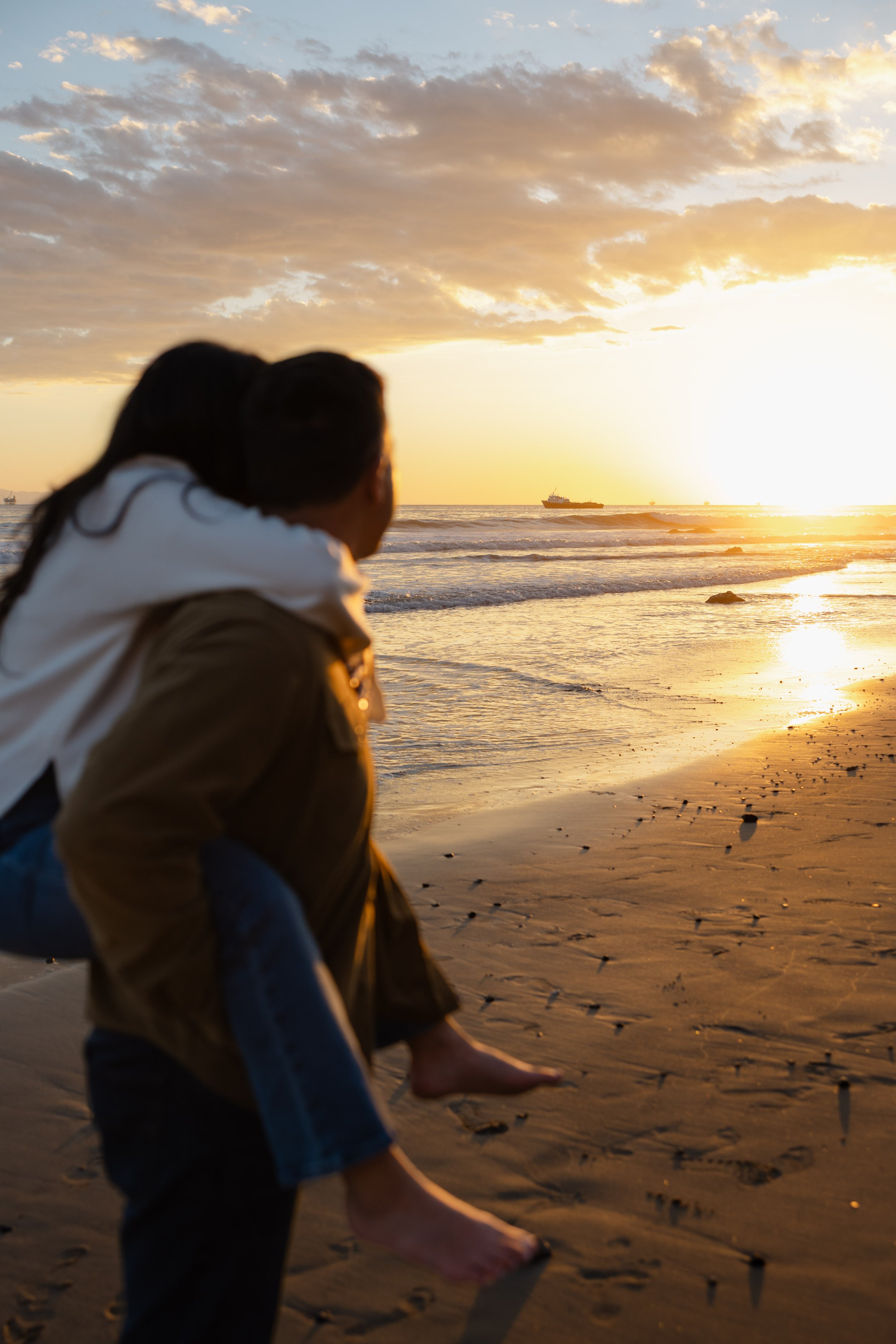 A couple at the beach during sunset, with the man carrying the woman on his back, both facing the horizon with boats in the distance.