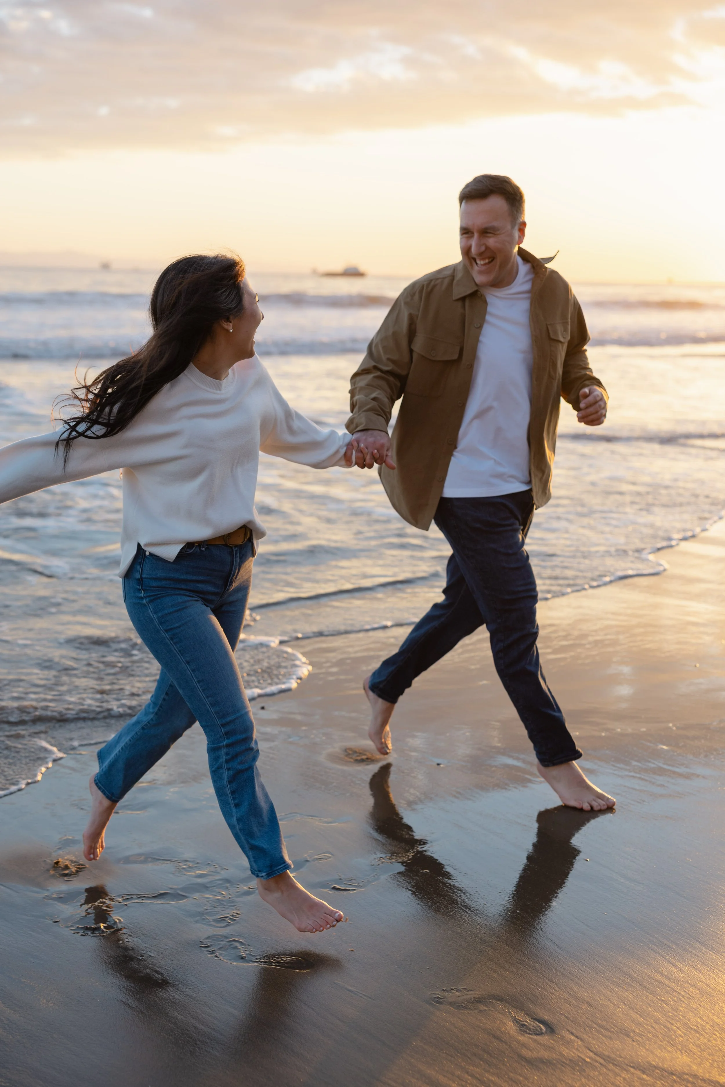 A man and woman are running and playing at the beach during sunset, holding hands and smiling.