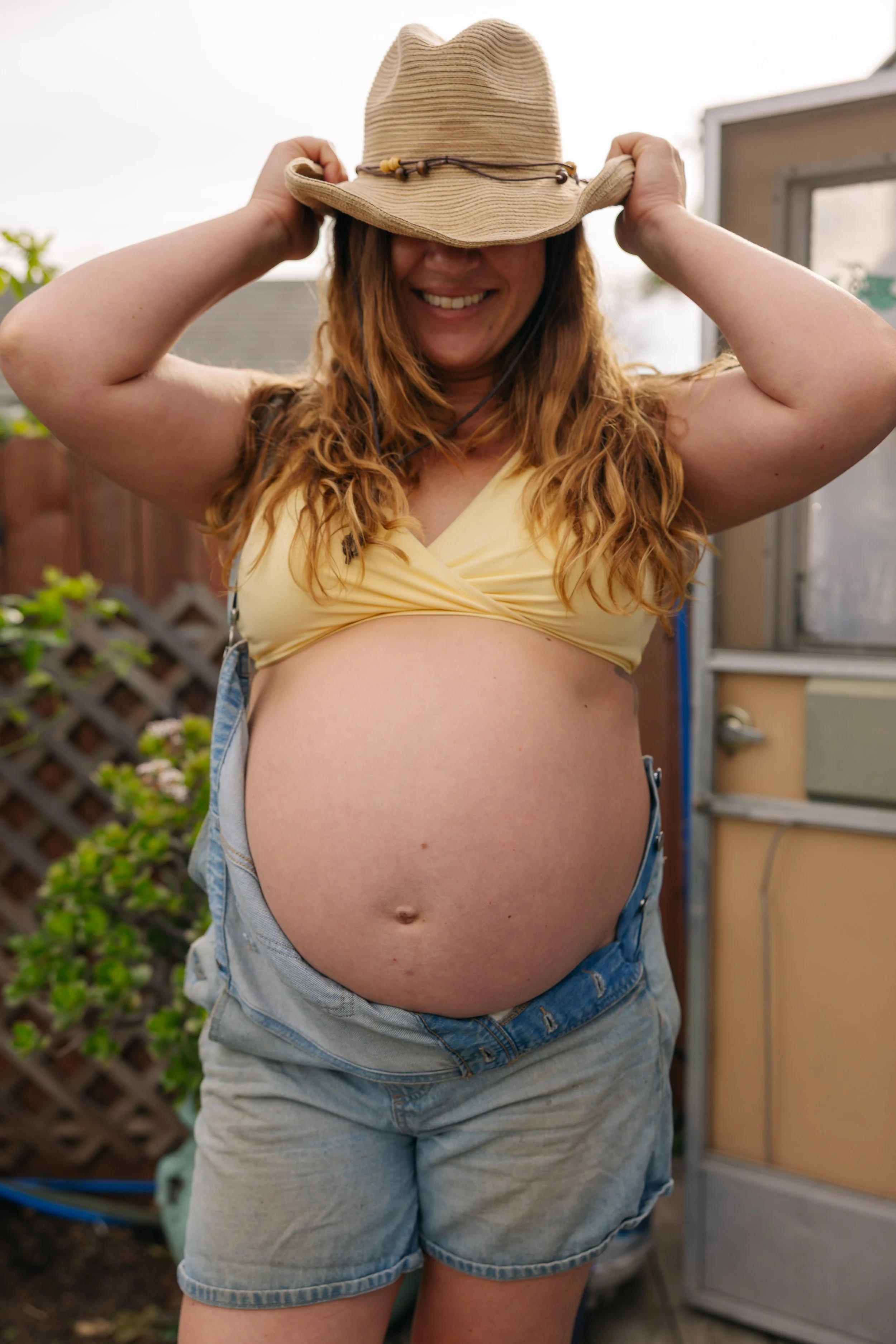 Smiling woman wearing a straw hat, a yellow crop top, and denim shorts, standing outdoors in a garden.