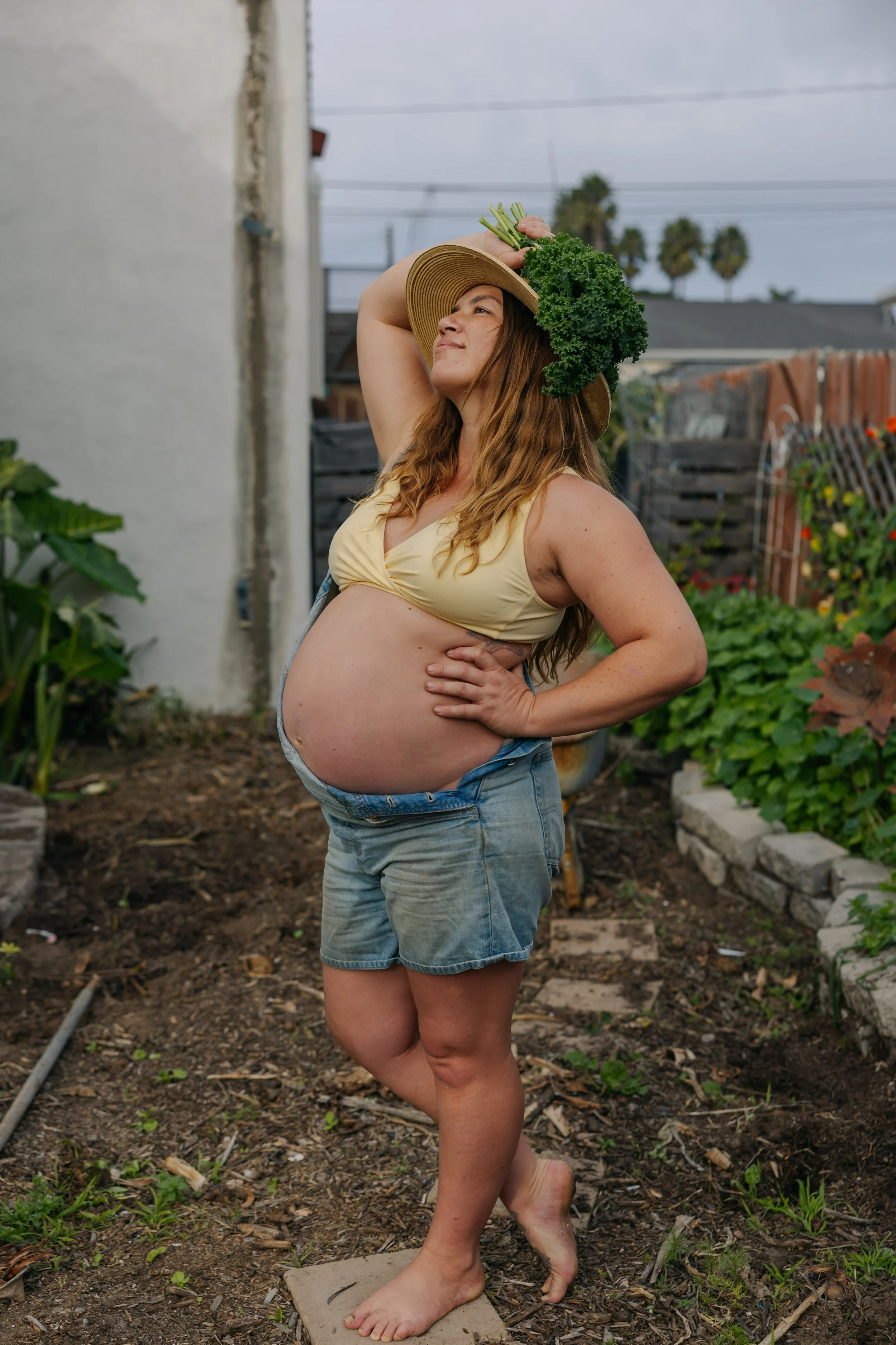 A pregnant woman stands outdoors in a garden, wearing denim shorts and a yellow tank top, holding a bunch of kale on a straw hat on her head, with her left hand on her hip and her right hand holding the kale hat, looking upwards.