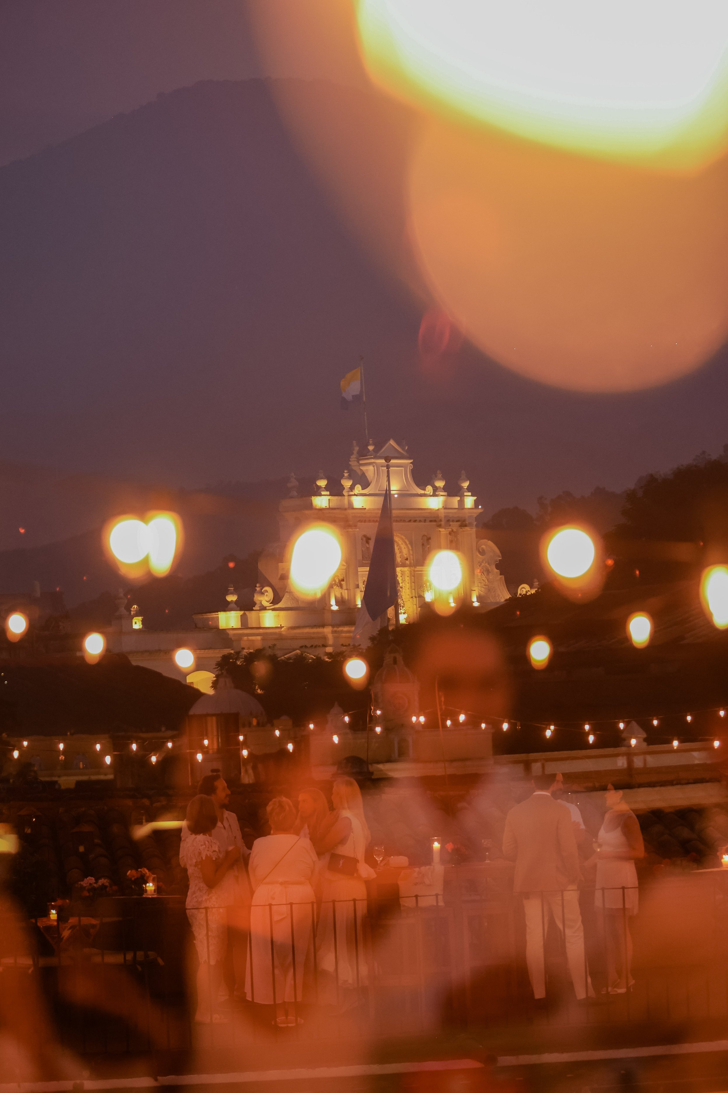 An evening scene of an outdoor gathering with people standing and chatting, viewed through a window with blurred orange and yellow lights. In the background, a grand, illuminated building with a flag on top, set against a dark mountain landscape.