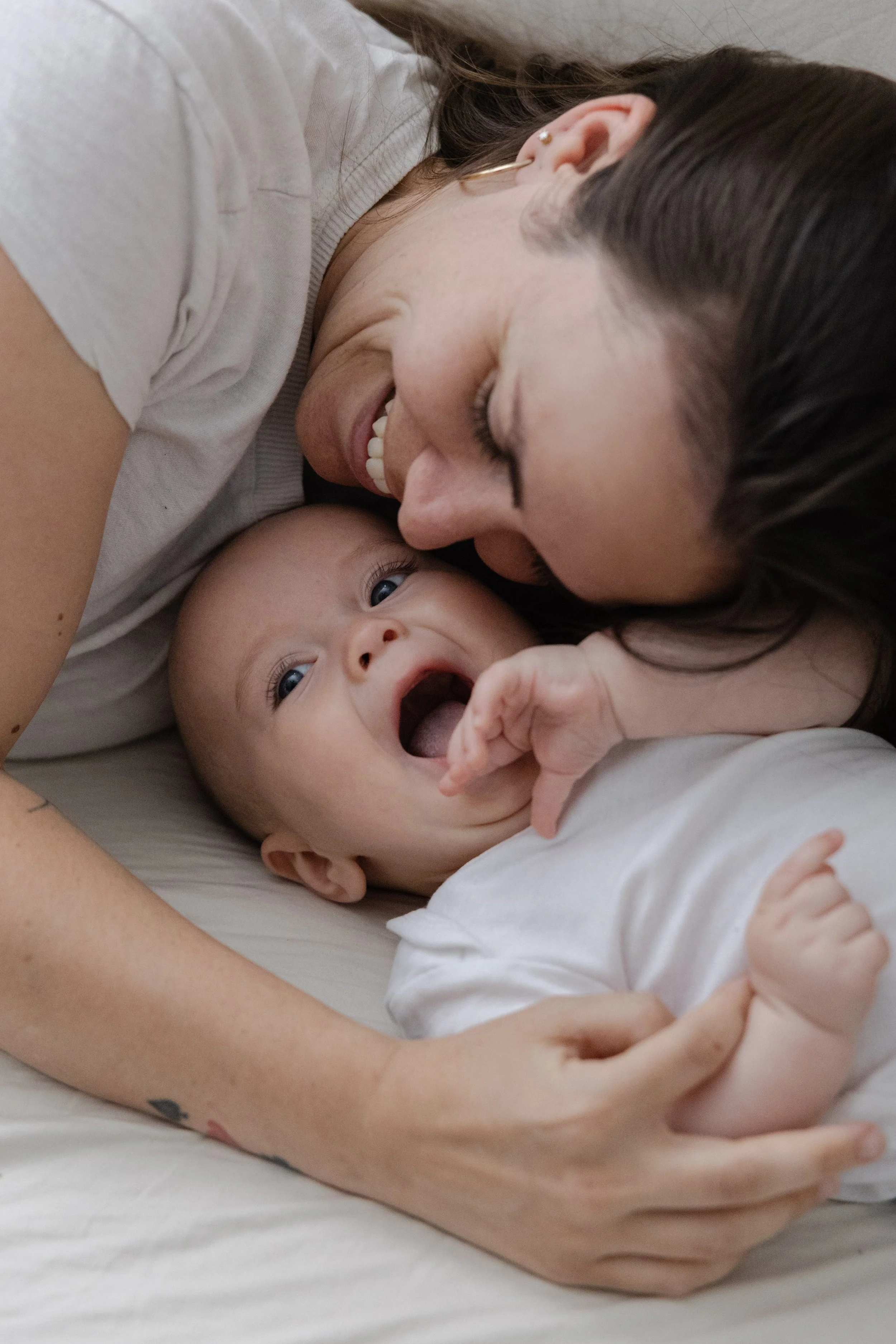 A woman and a baby lying on a bed, close together, smiling and playing. The woman is touching the baby, who has an open mouth and is reaching with his hand.