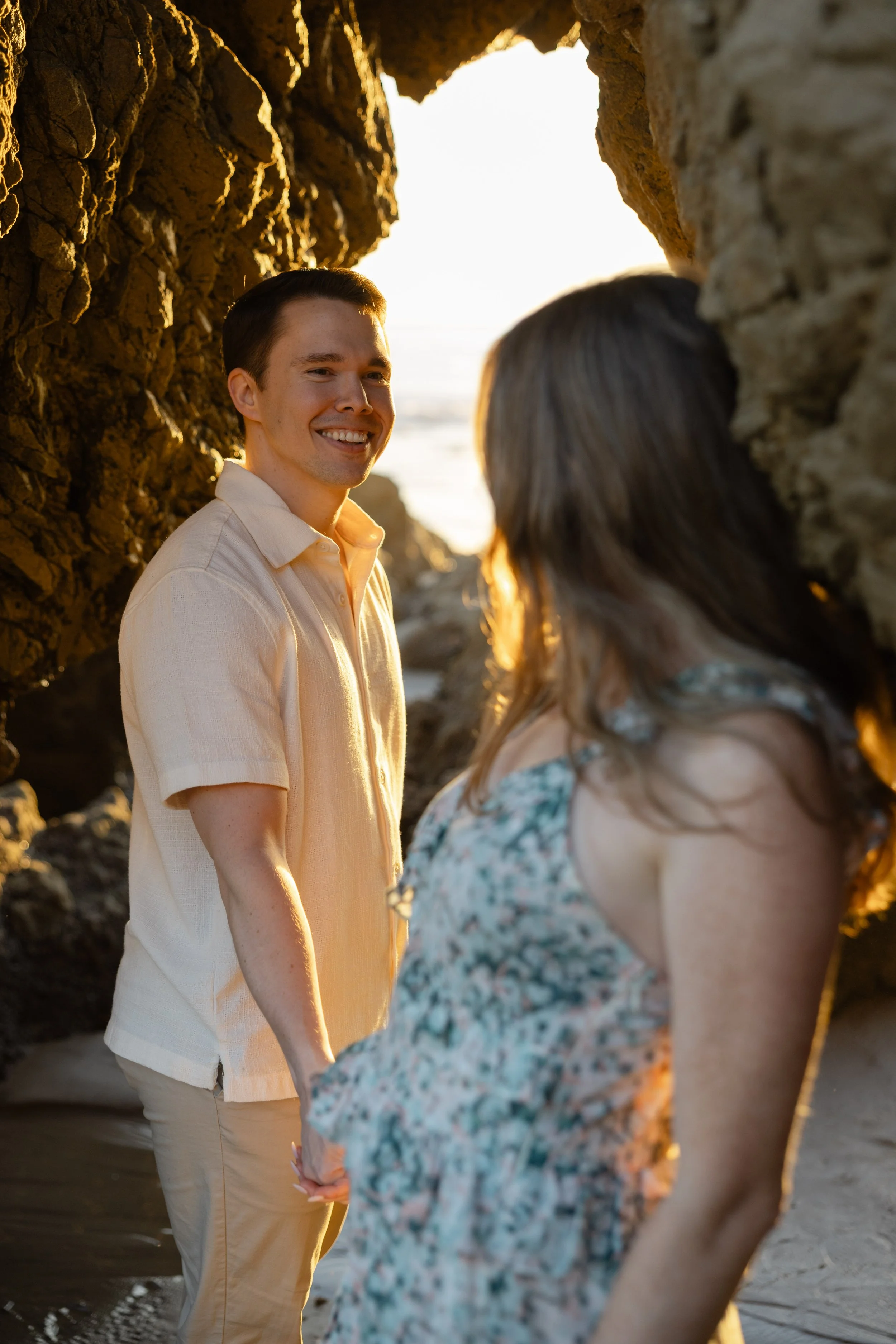A young man and woman holding hands and standing between large rocks at the beach during sunset, with the ocean in the background.