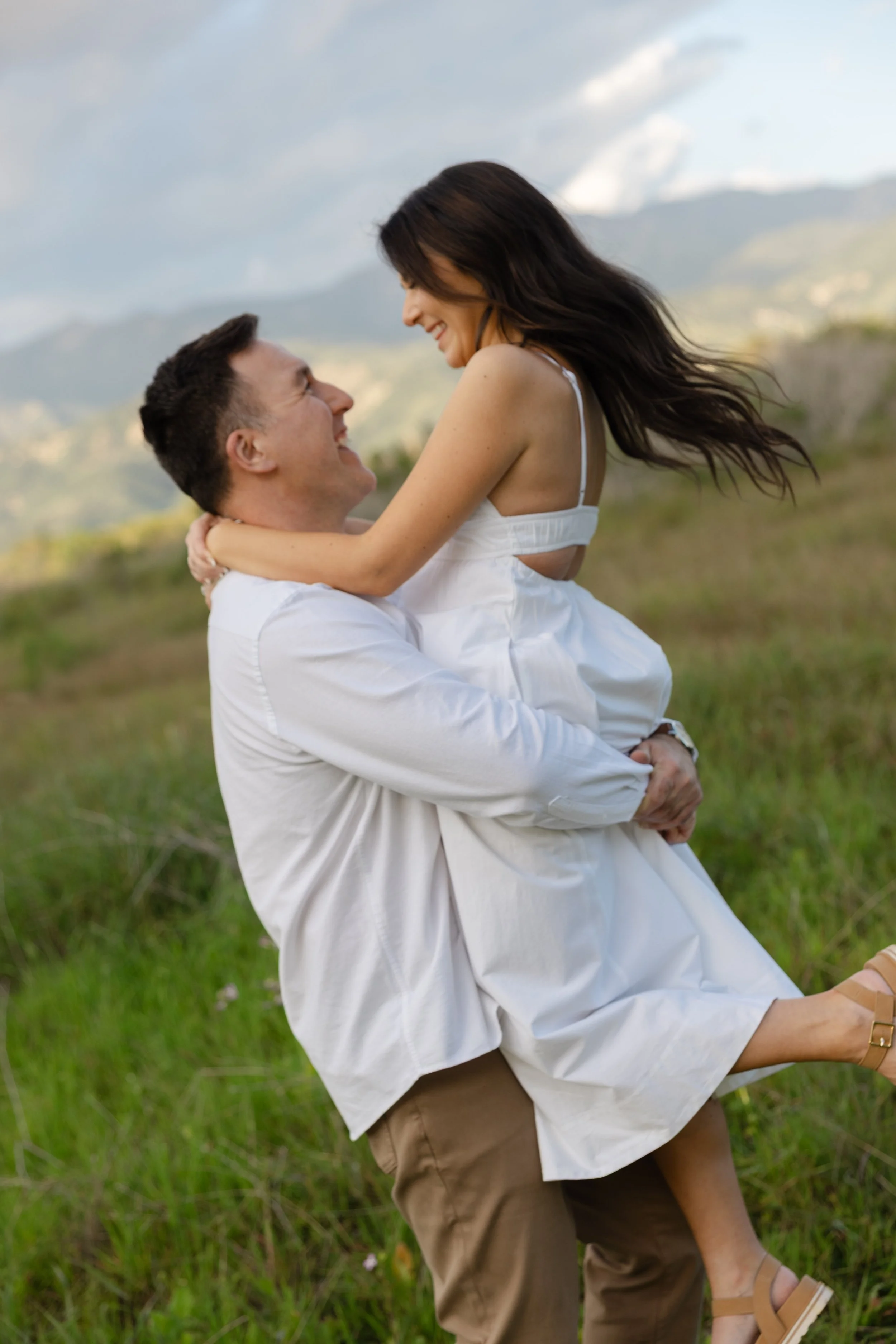 A man lifting a woman in a white dress outdoors in a grassy field with mountains in the background, both smiling and looking at each other.