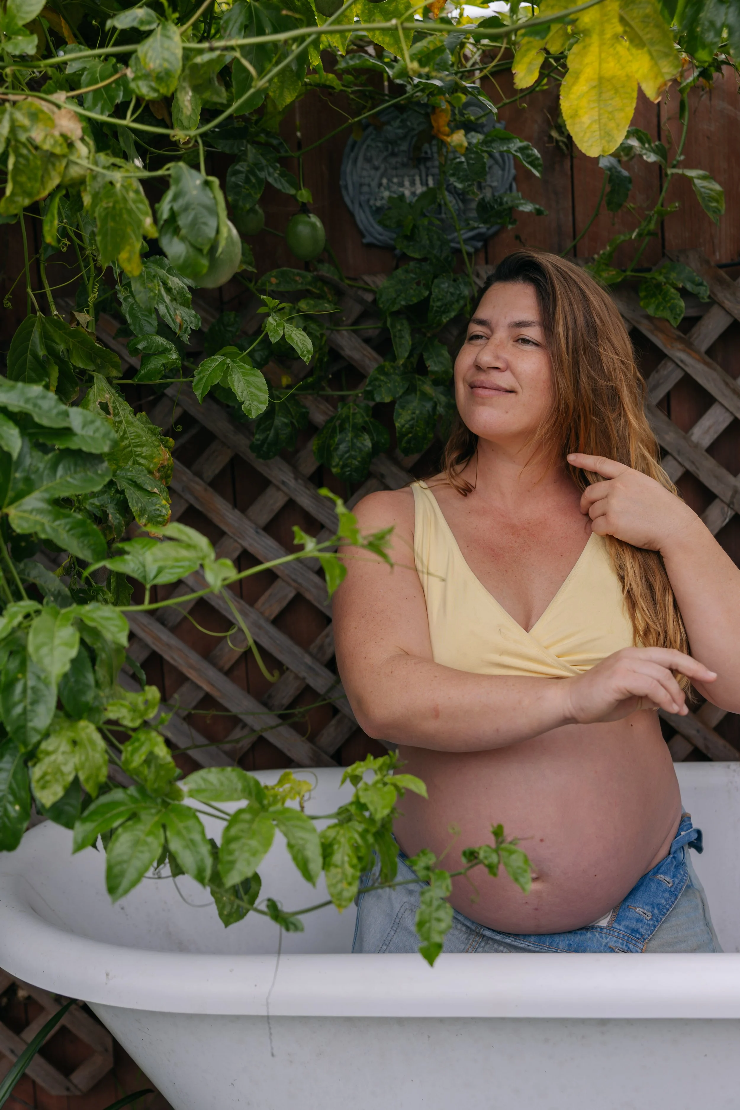 Pregnant woman in a yellow top sitting in a bathtub outdoors, surrounded by green plants and a wooden fence.