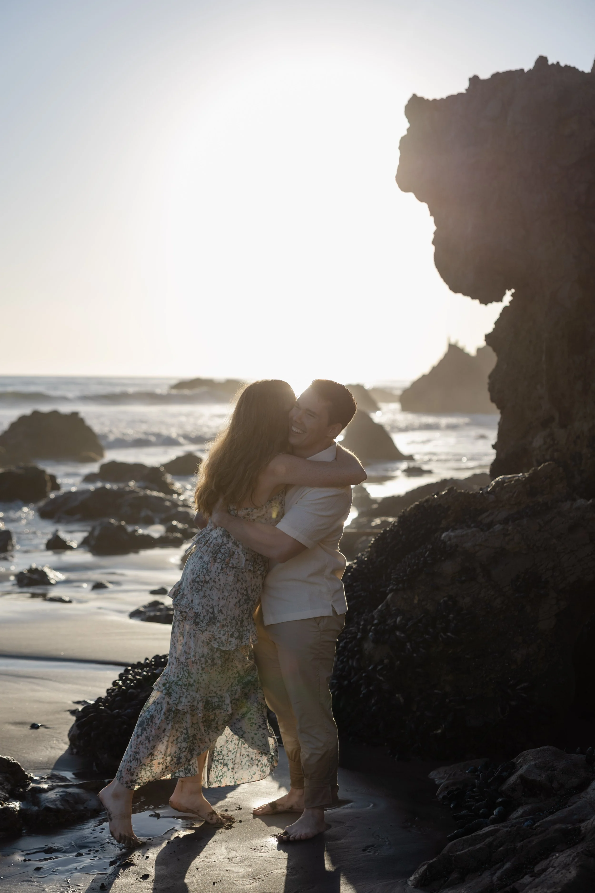 A couple hugging on a beach at sunset with rocks and waves in the background.