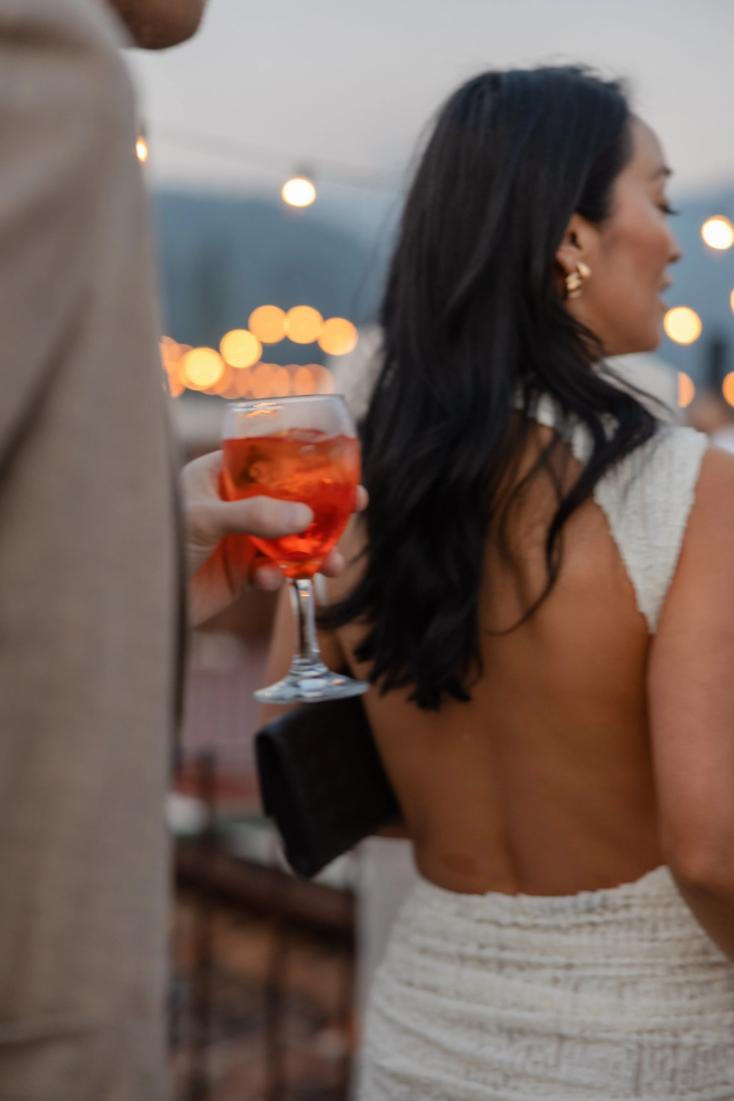 A woman with dark hair and jewelry standing outdoors at sunset, holding a glass of red-orange drink with ice, while another person wearing a beige jacket is nearby. String lights illuminate the background.