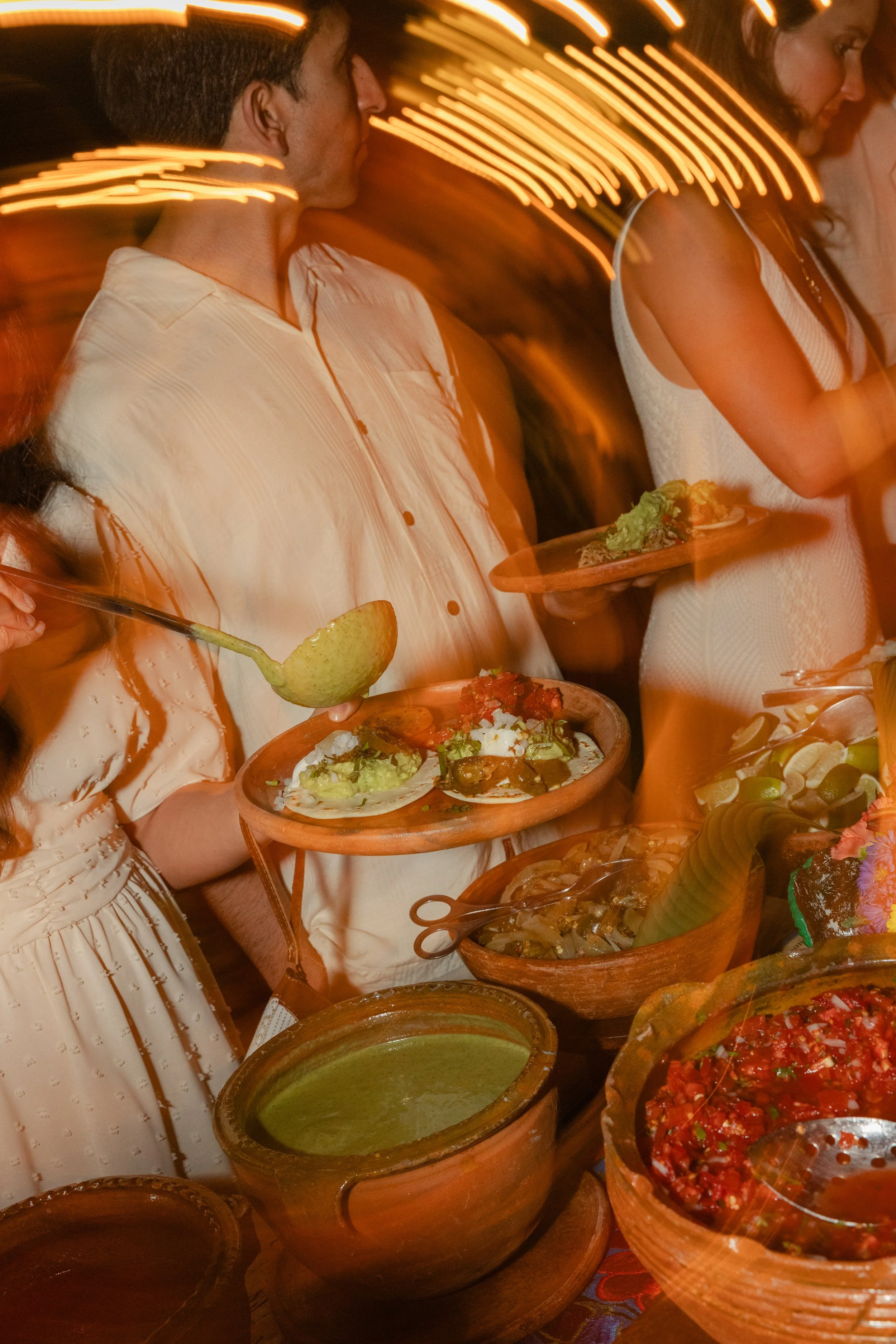 People standing in line at a buffet table with various Mexican foods, including green salsa, guacamole, and chili, with decorative warm lighting.