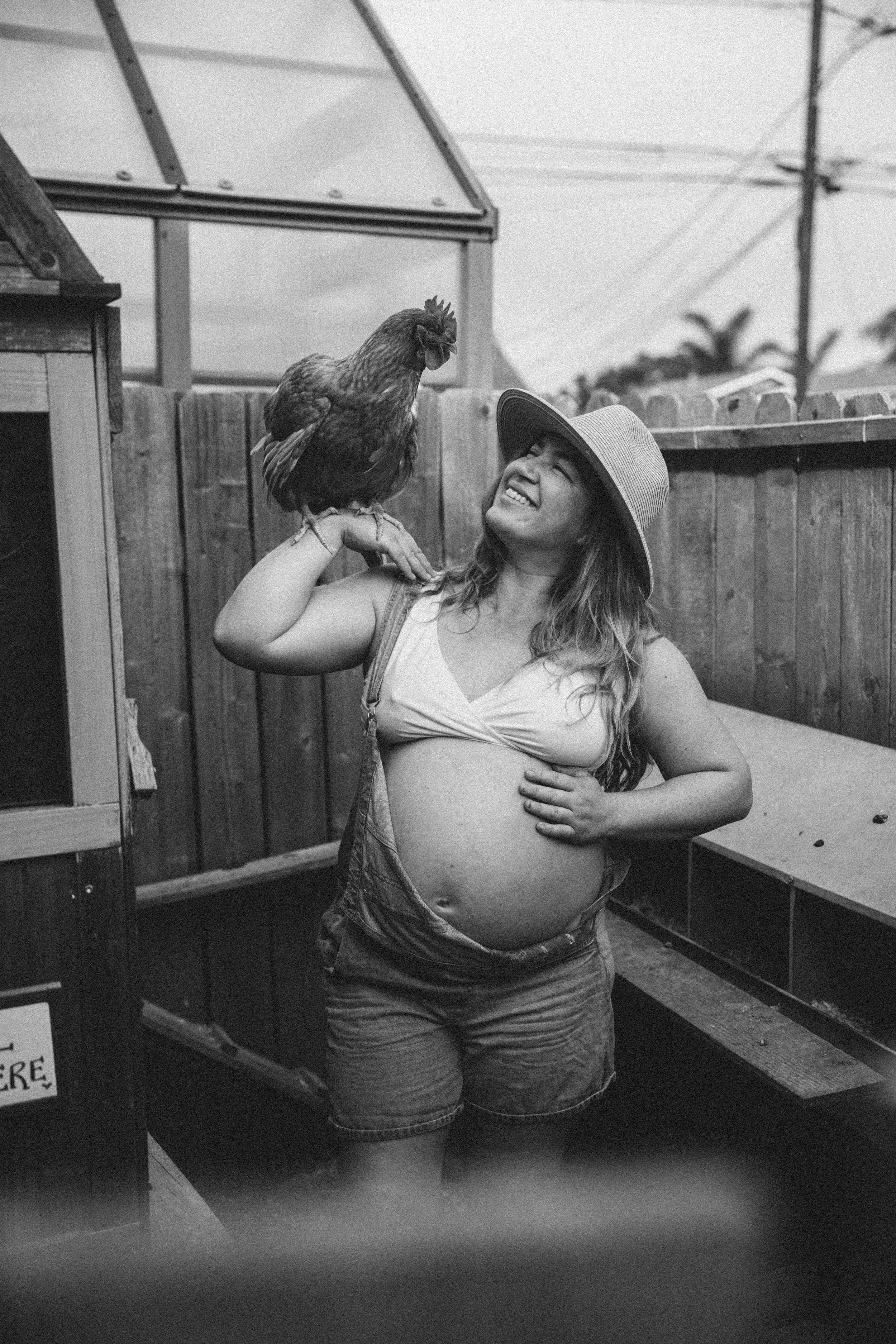 A pregnant woman smiling and looking at a chicken perched on her shoulder in a backyard enclosed by a wooden fence, with a greenhouse and power lines in the background.