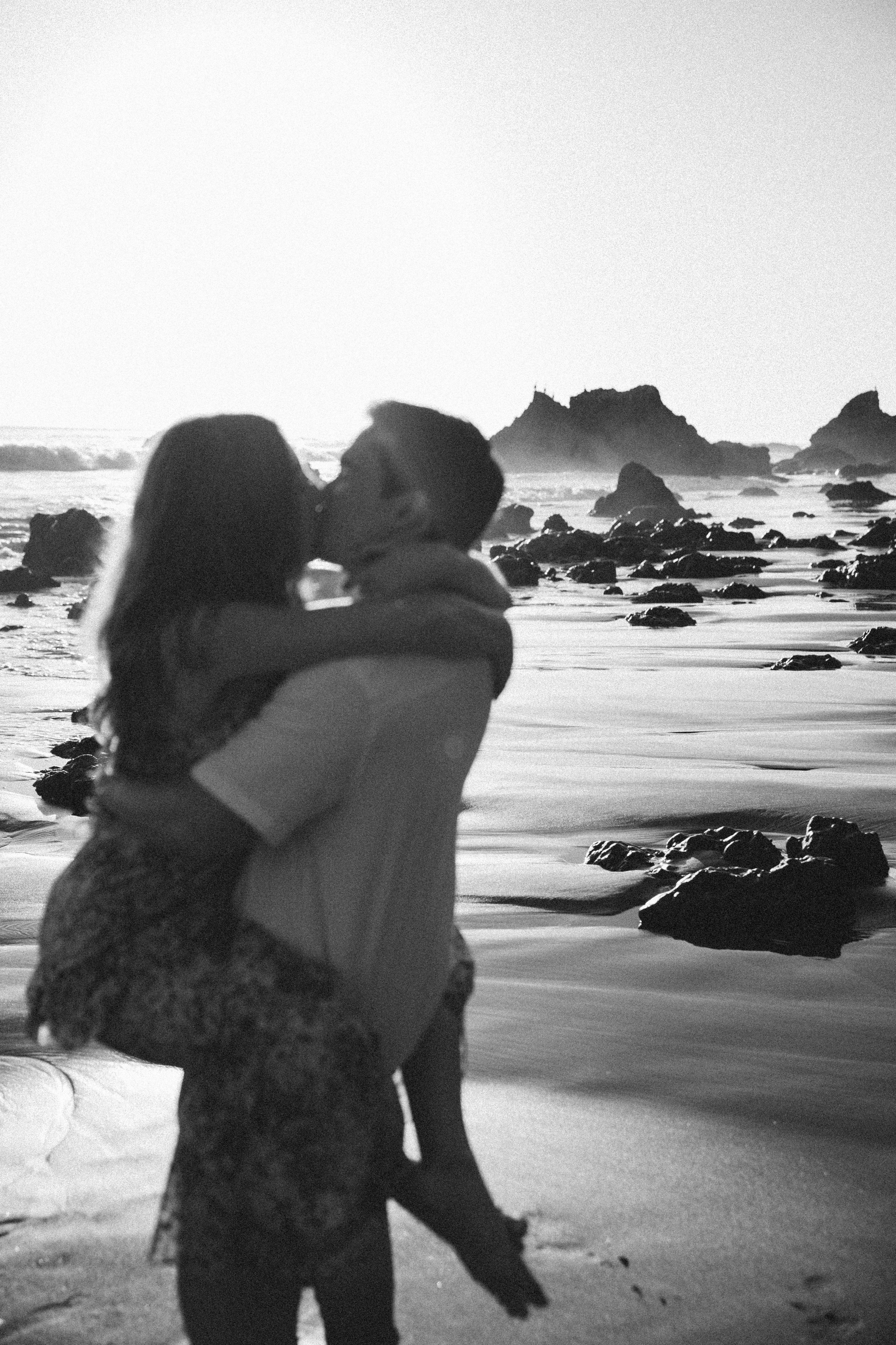 A black-and-white photo of a couple kissing on a beach, with rocks and ocean waves in the background.