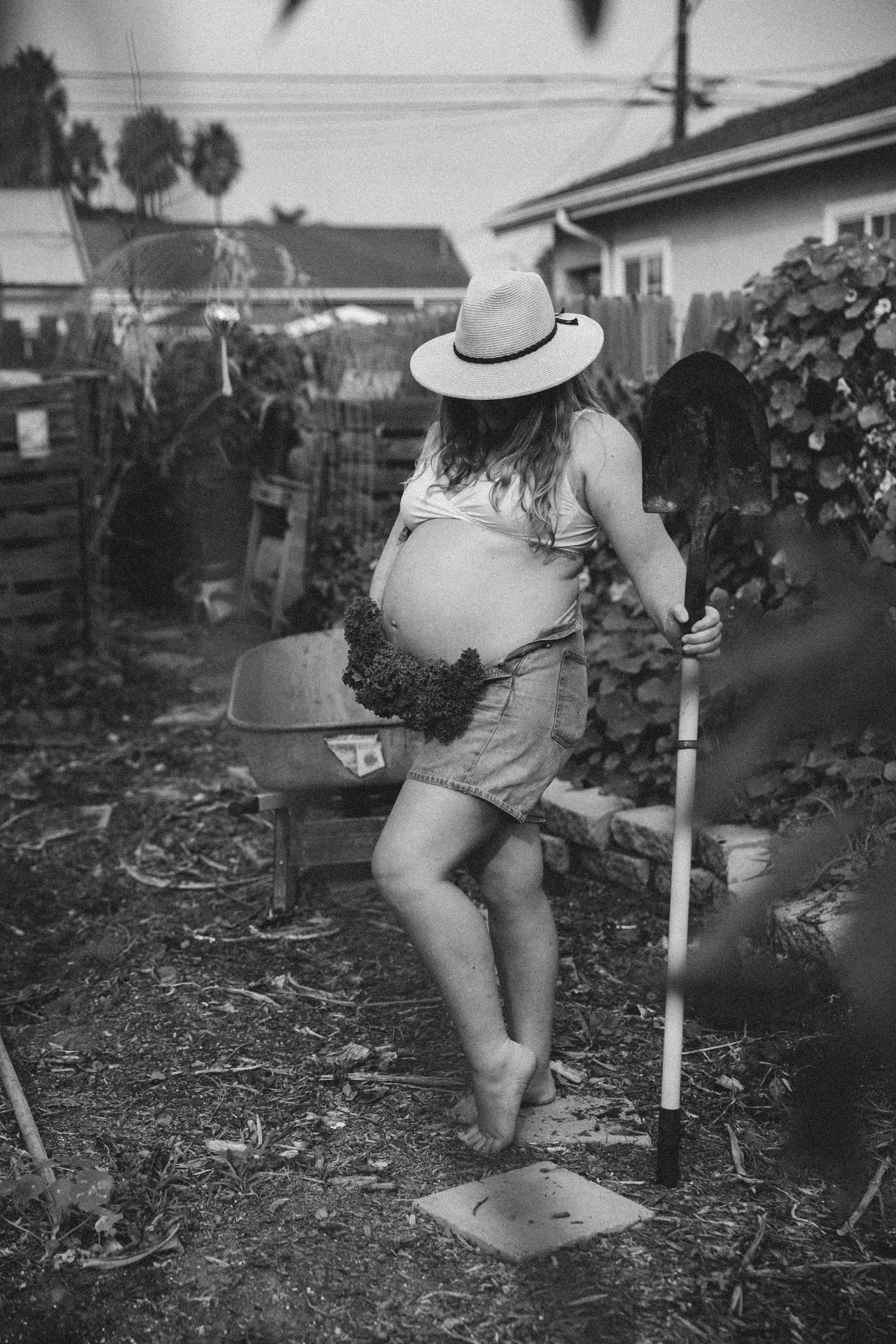 A person with long hair wearing a wide-brimmed hat, shorts, and only a piece of cloth covering their abdomen, holding a shovel, standing barefoot on dirt in a backyard with plants and gardening tools.