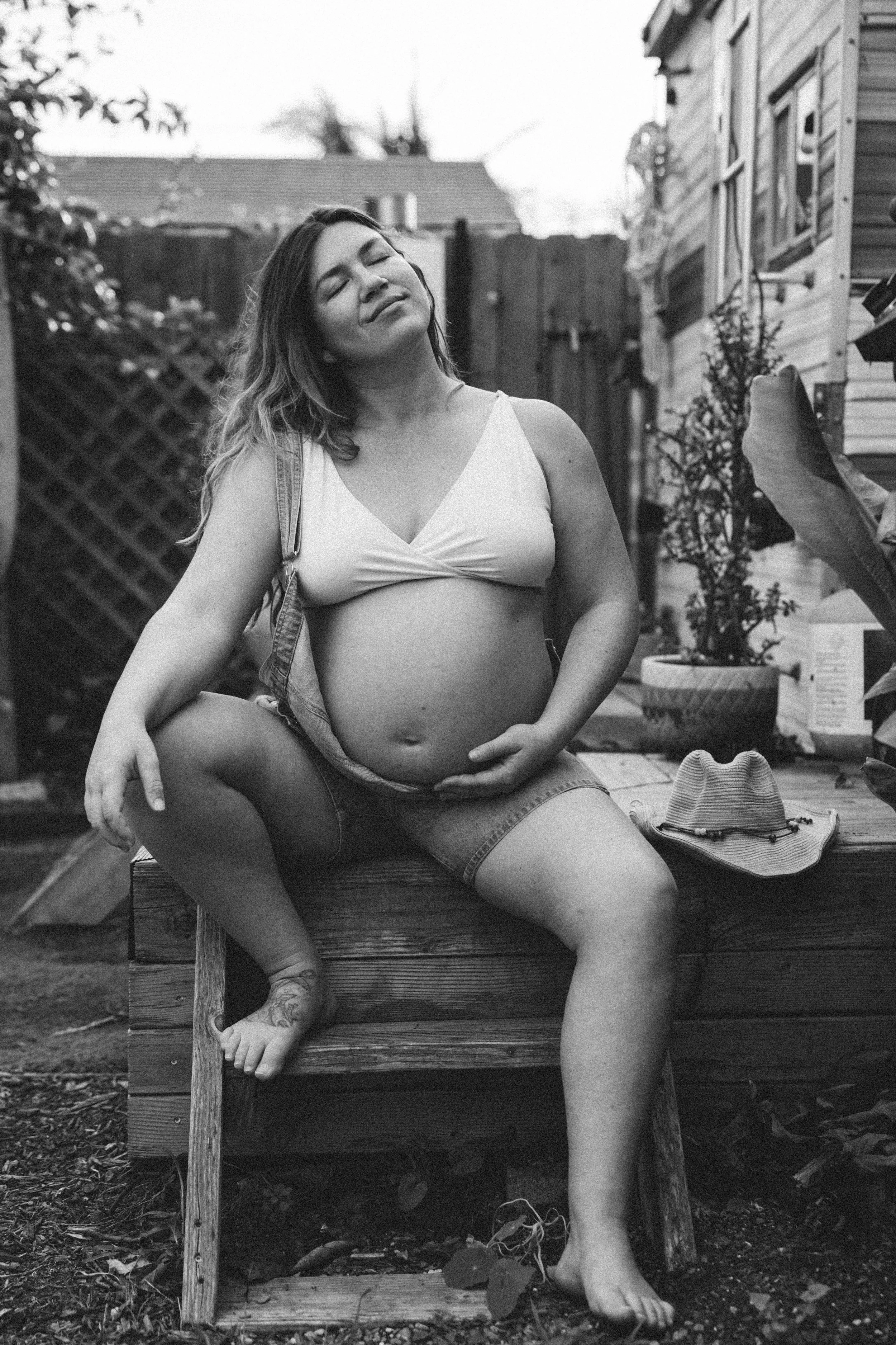 A pregnant woman sitting on a wooden bench outdoors, with a relaxed expression, wearing a tank top and shorts, with a hat and jewelry beside her.
