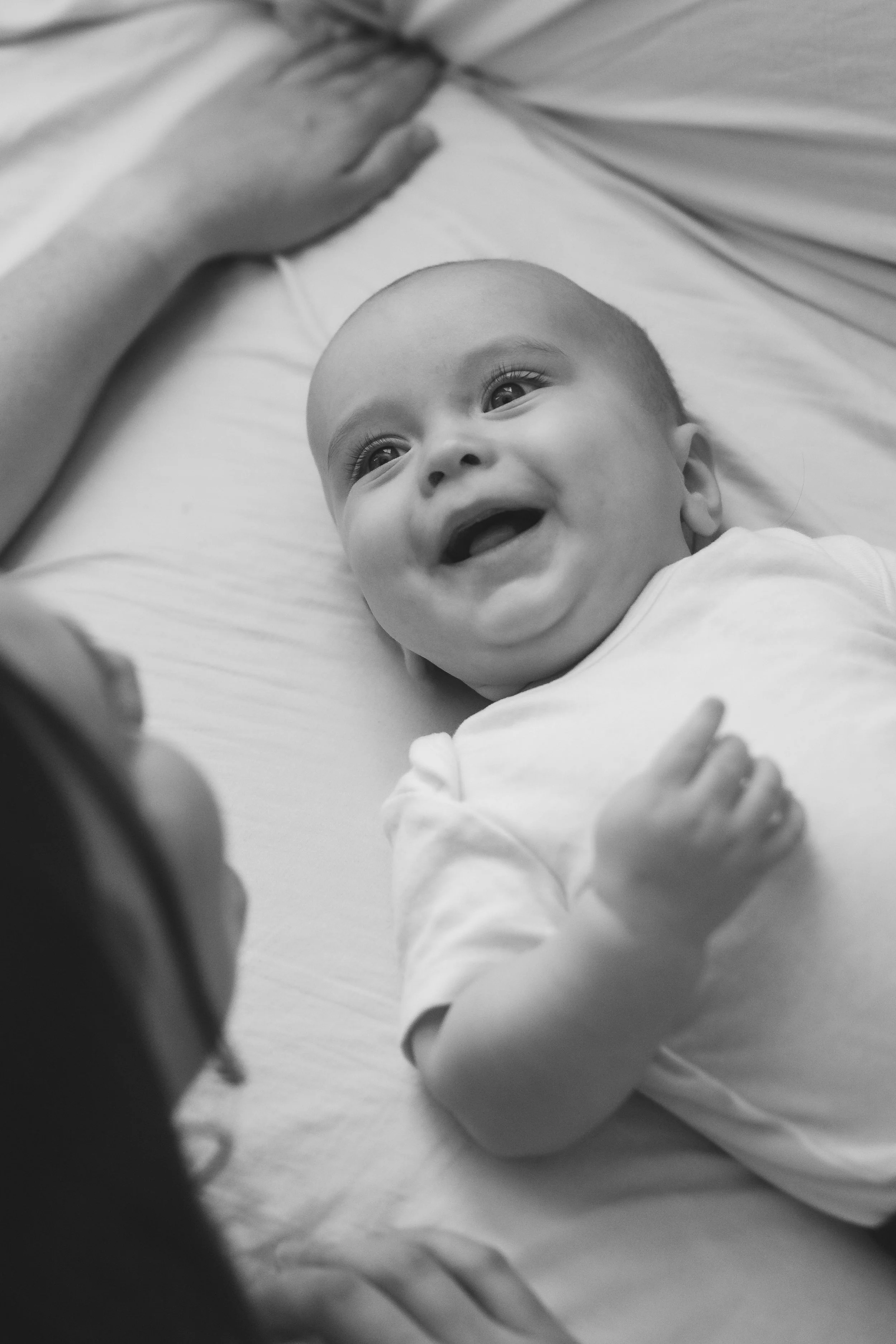 A smiling baby lying on a bed, looking up at a person holding a camera, with an adult's arm visible resting nearby.