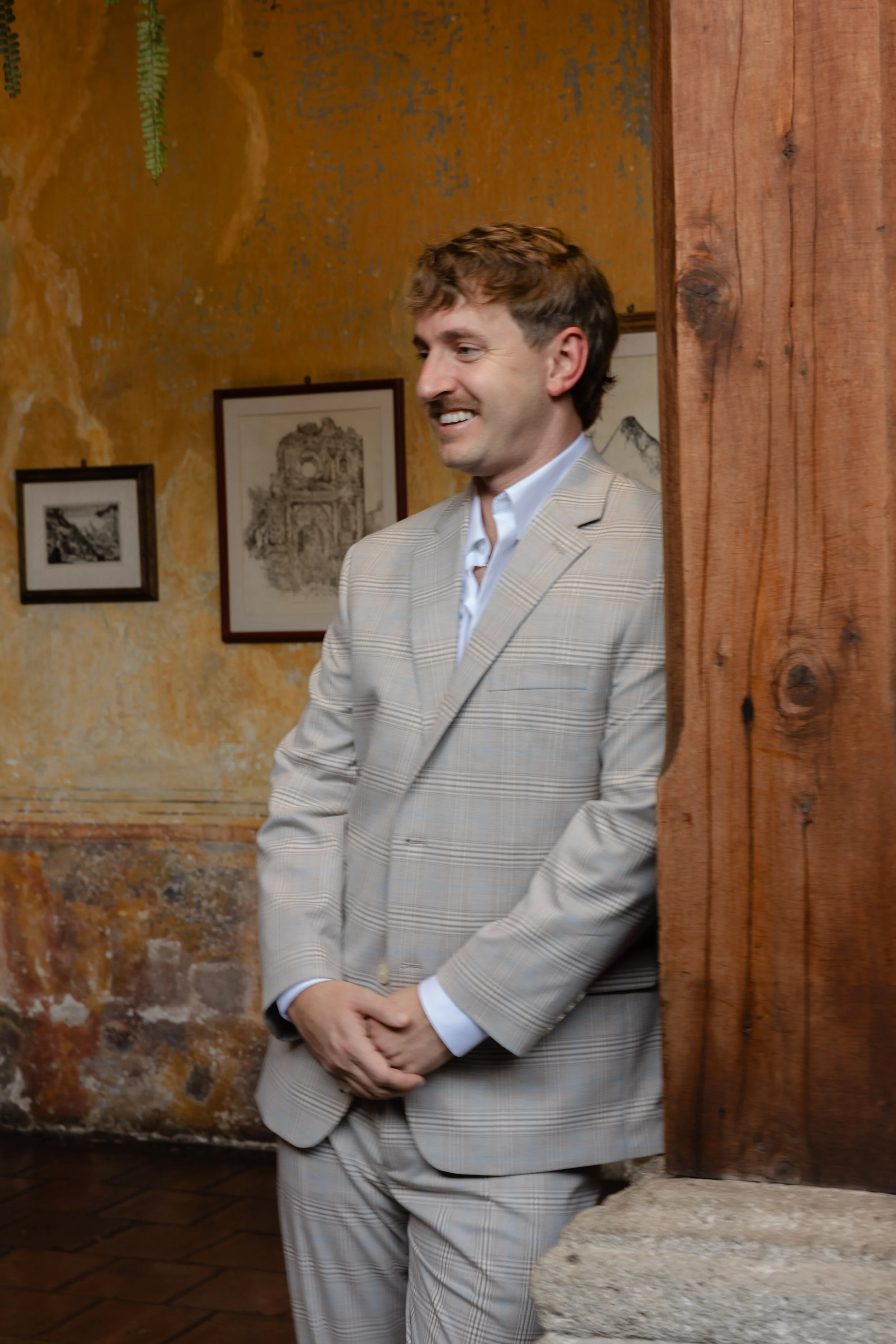 A man in a light-colored checkered suit smiling and leaning against a wooden column in a warmly decorated room.