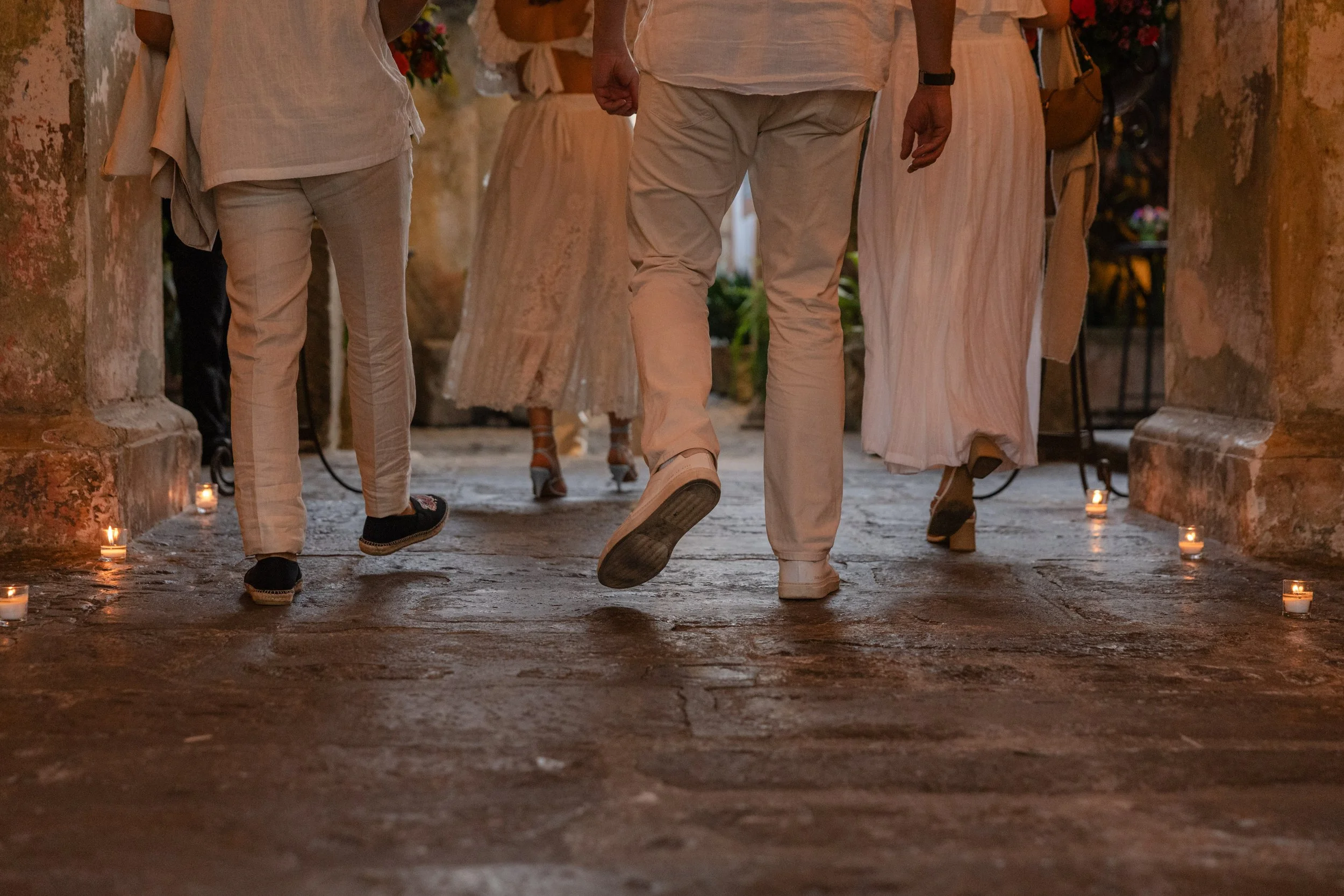 People walking in a candlelit corridor with candles on the ground, wearing light-colored clothing, possibly at an outdoor event or celebration.